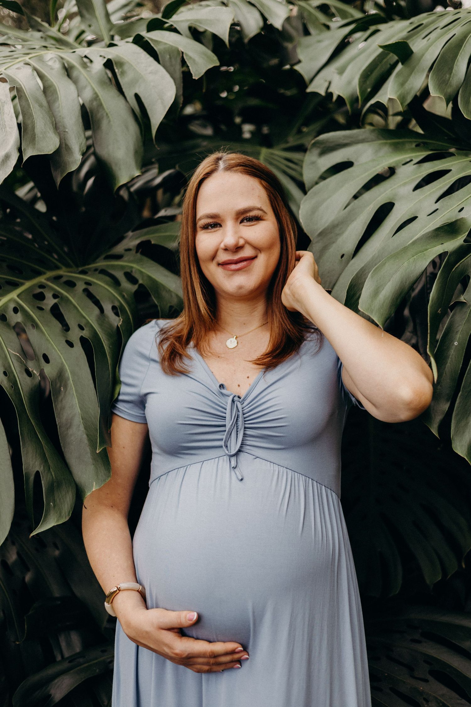 A pregnant woman in a blue dress poses against a backdrop of large monstera leaves in a maternity photo series.