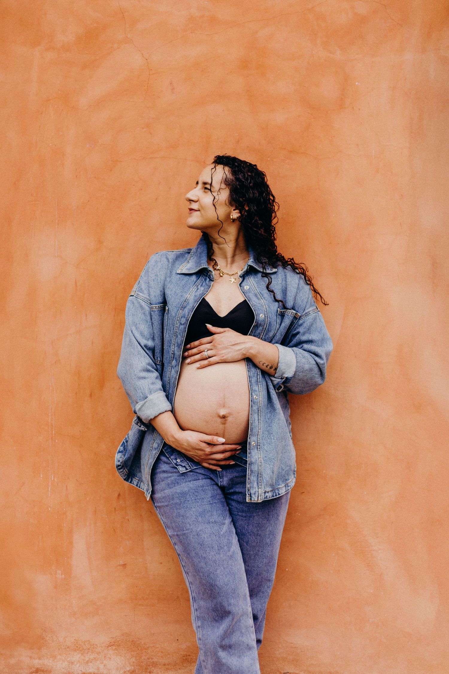 Pregnant woman in denim jacket and jeans poses against orange wall while holding her belly.