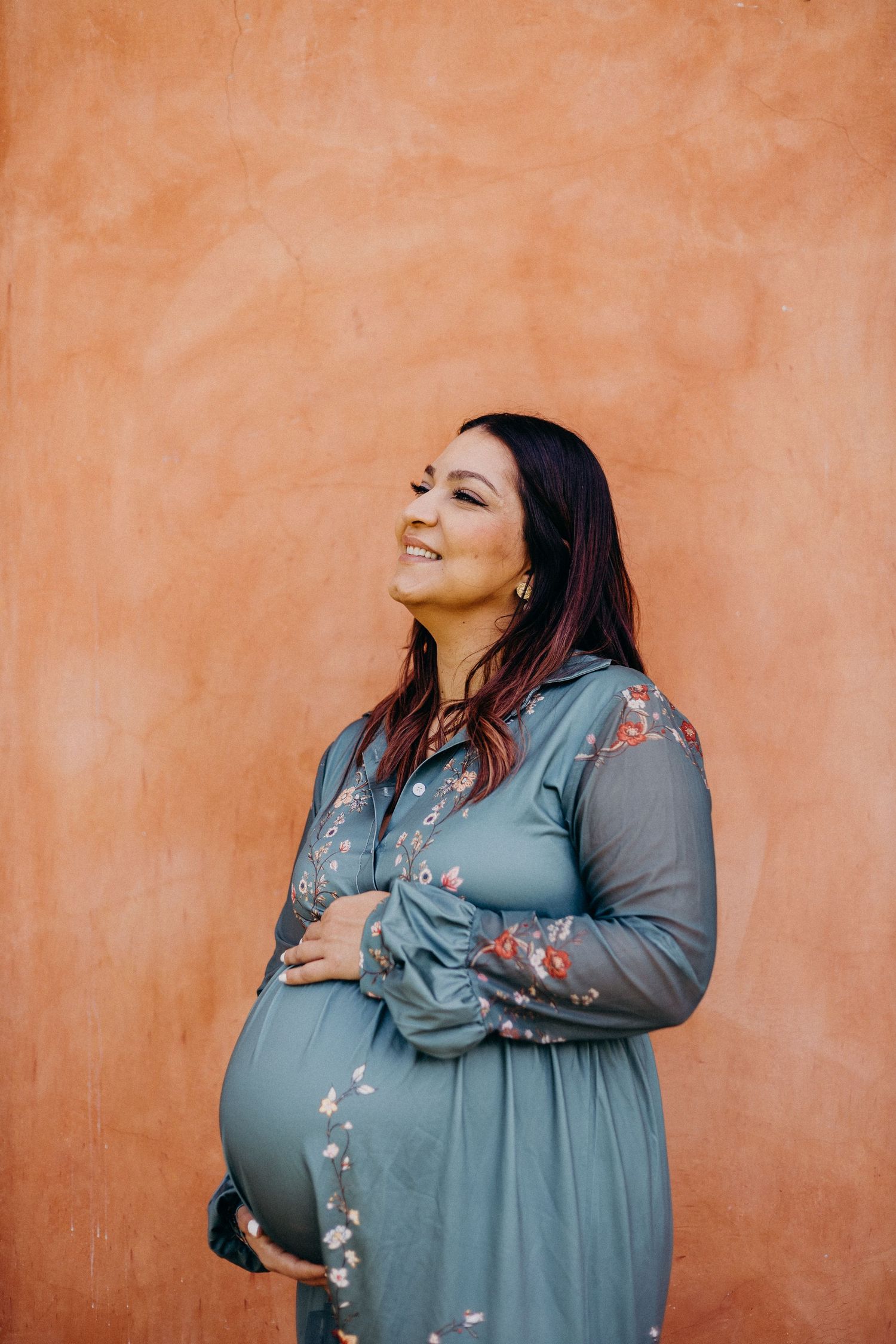 A pregnant woman in a blue floral dress smiles while standing against a terracotta colored wall.