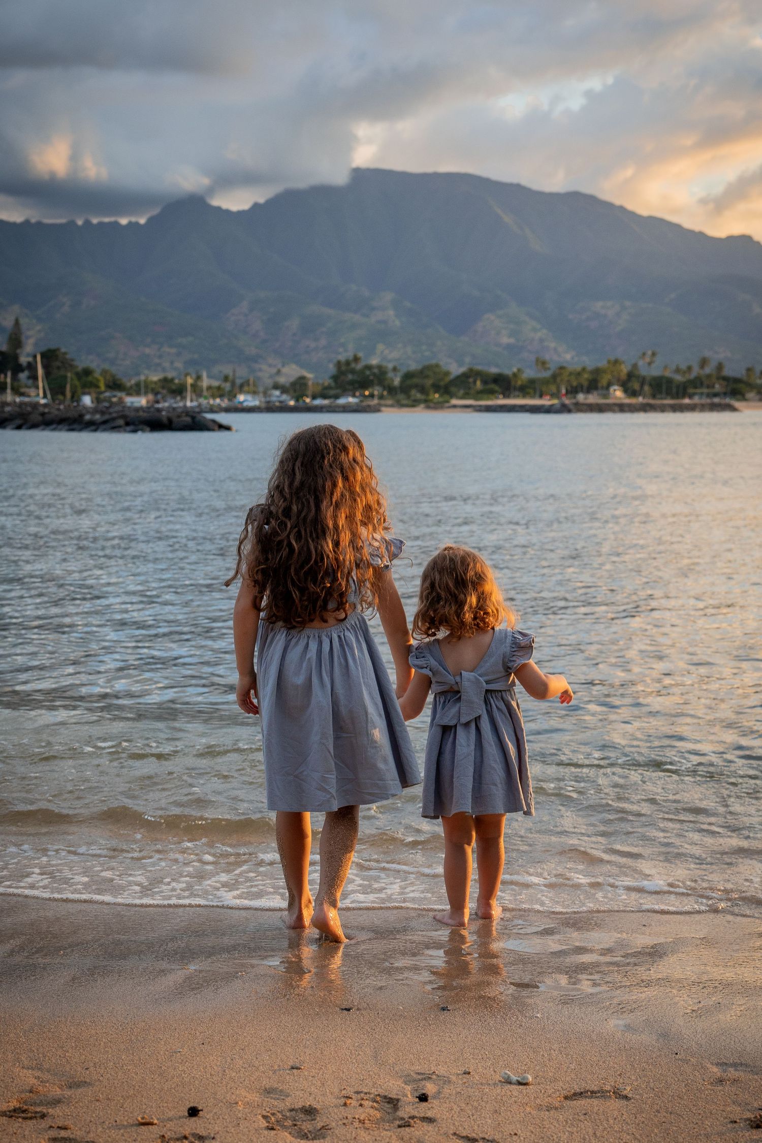 Two people in matching dresses stand at the ocean's edge during sunset with mountains in background.