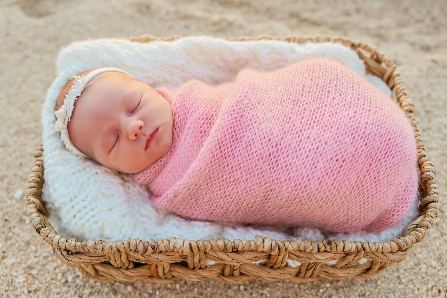 A sleeping newborn wrapped in pink swaddle rests in a woven basket.