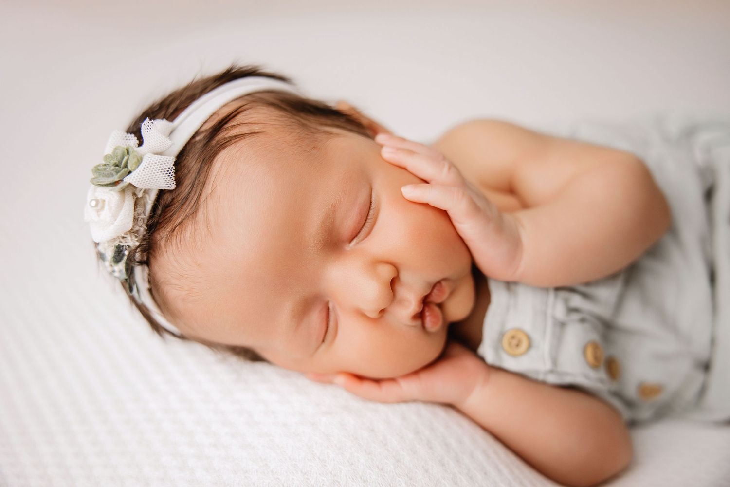 Sleeping newborn wearing floral headband and grey outfit on white textured surface.