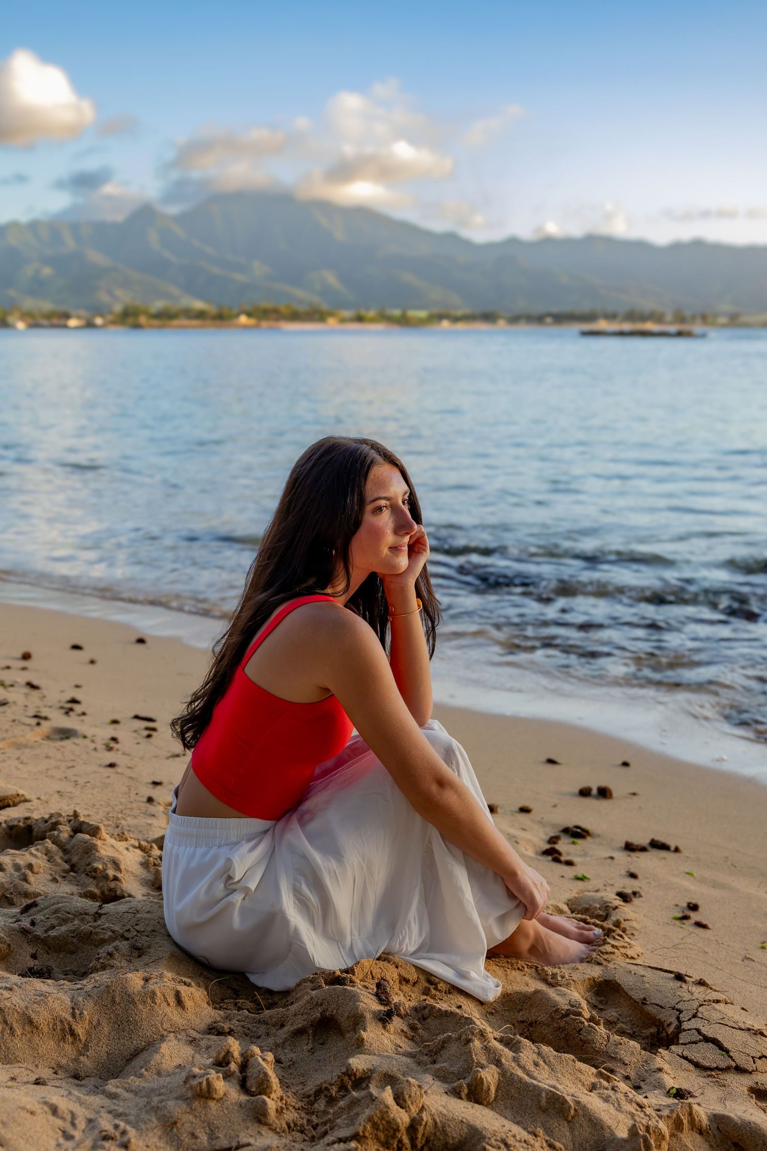 A woman in red sits peacefully on a beach with mountains and ocean in the background.