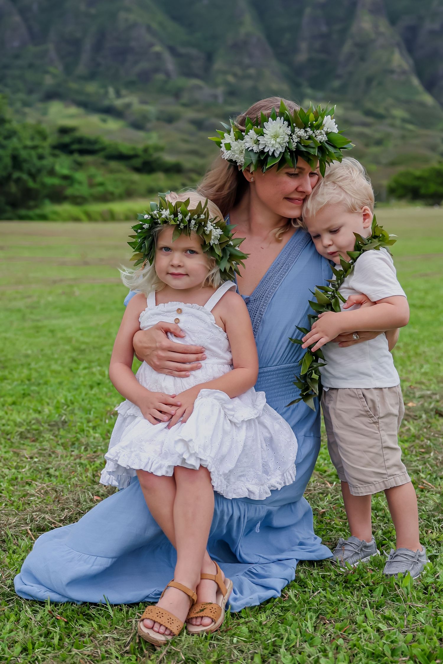 Family wearing flower leis pose together on grassy field with dramatic mountain landscape.