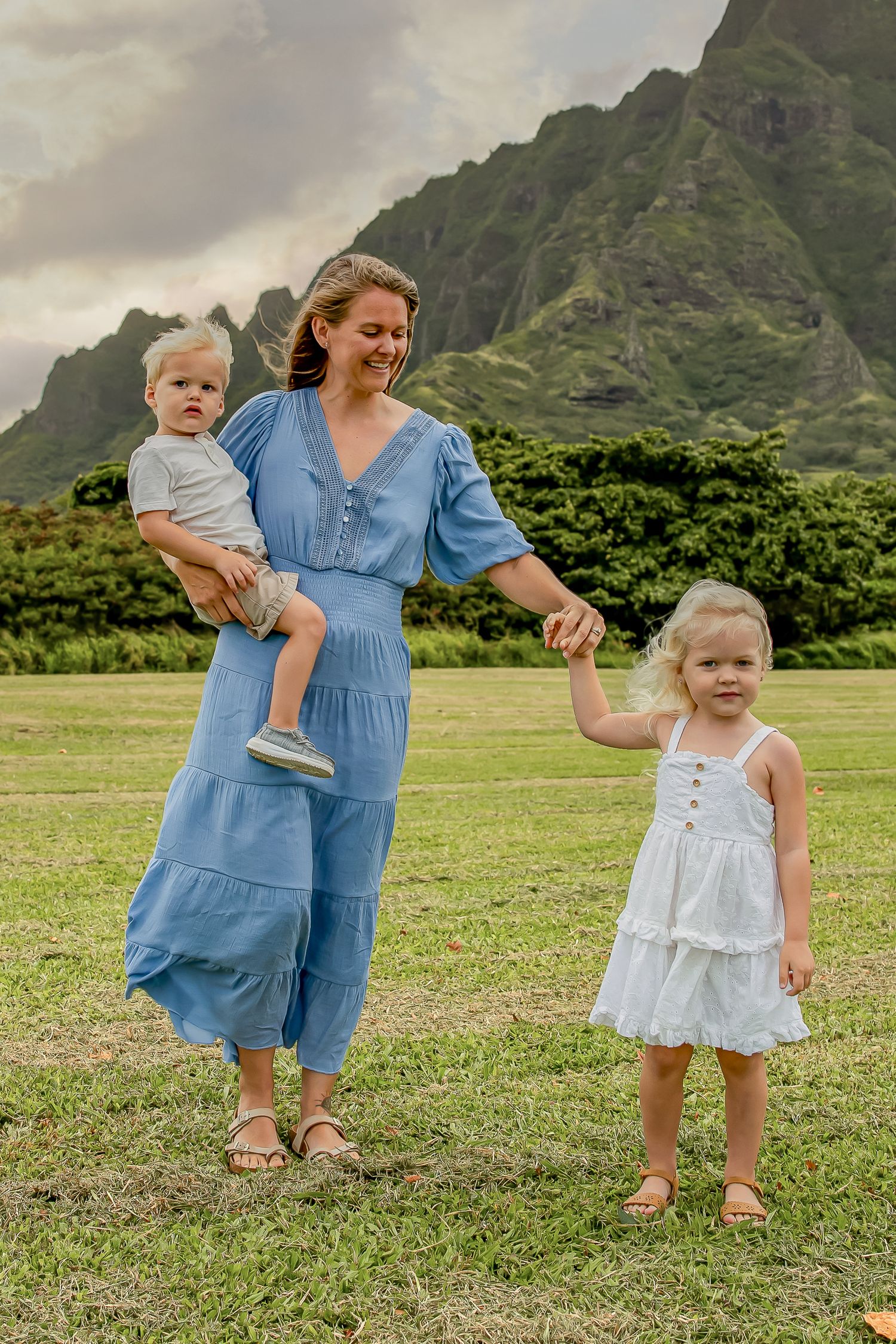 A family stands together in a grassy field with dramatic mountain peaks rising in the background in Hawaii.