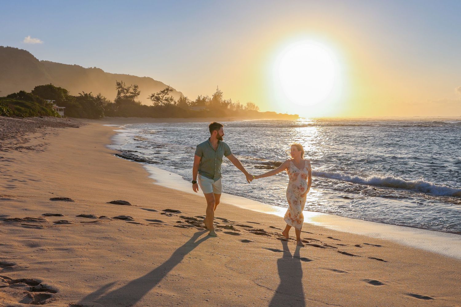 Couple walking hand in hand on sandy beach during dramatic sunset with mountain backdrop.