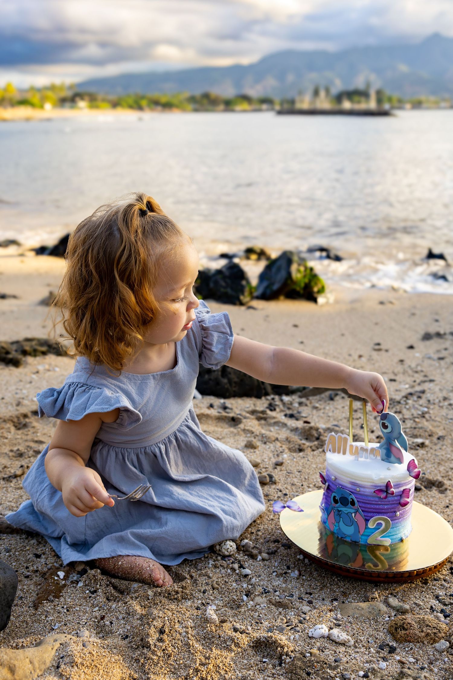 Young child in blue dress playing with a decorative cake on a sandy beach with mountains in the background.
