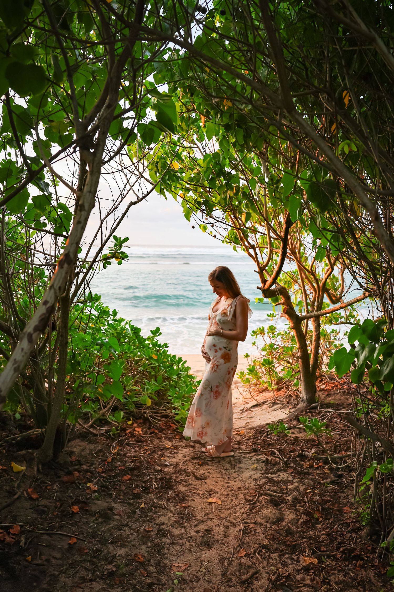 A person in a flowy dress stands on a scenic coastal trail framed by lush trees overlooking the ocean.