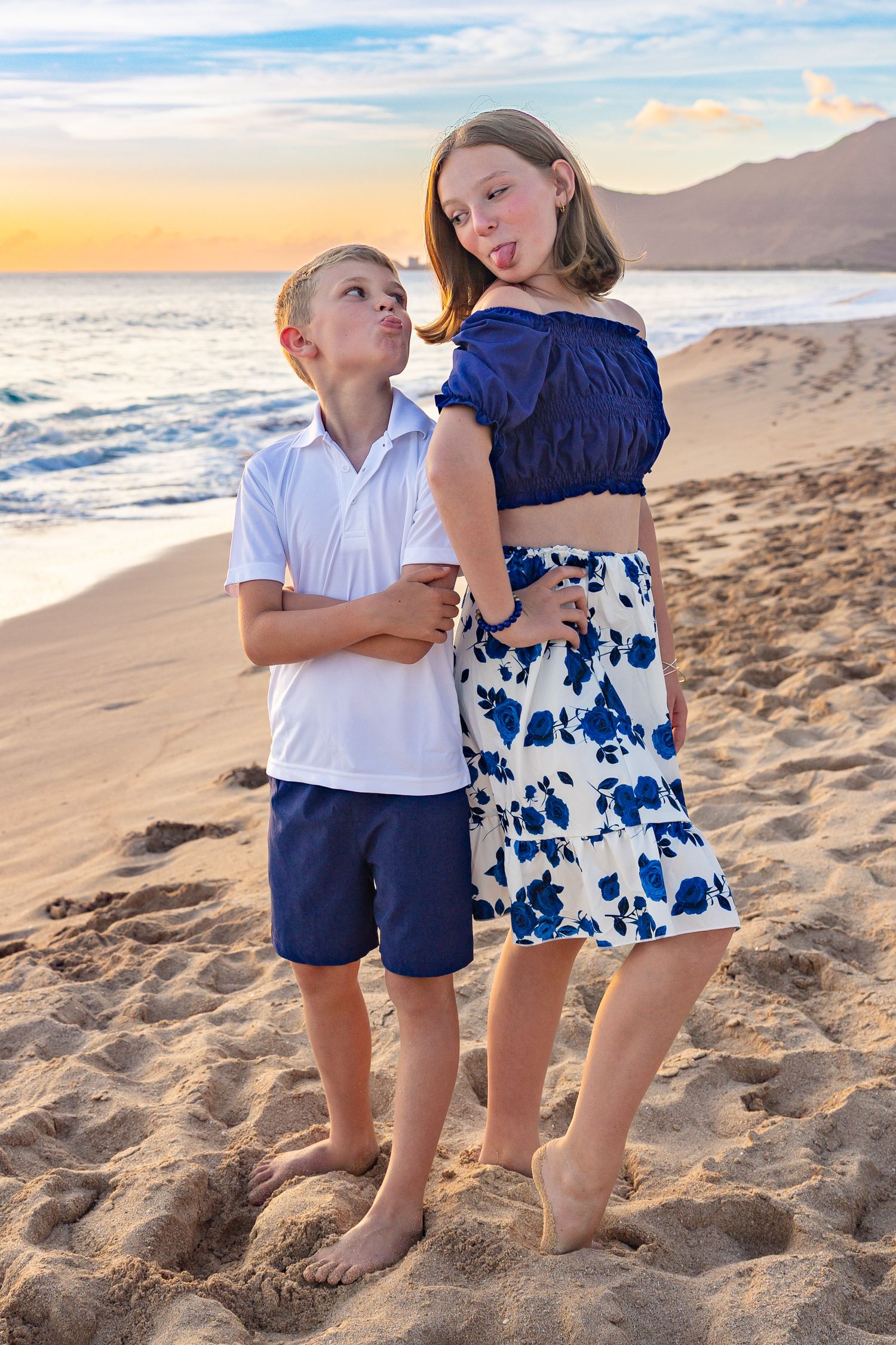 Two children posing together at sunset on sandy beach with mountains in background.