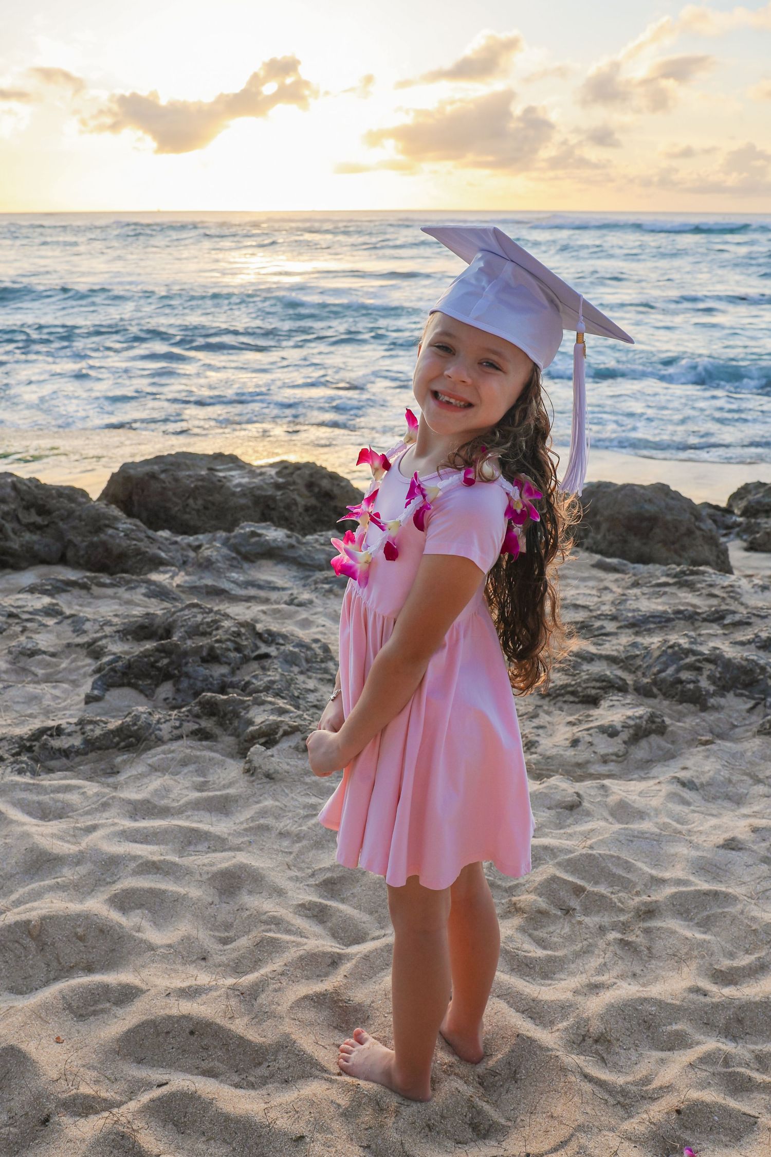 A young graduate in pink dress and lei poses on beach at sunset.