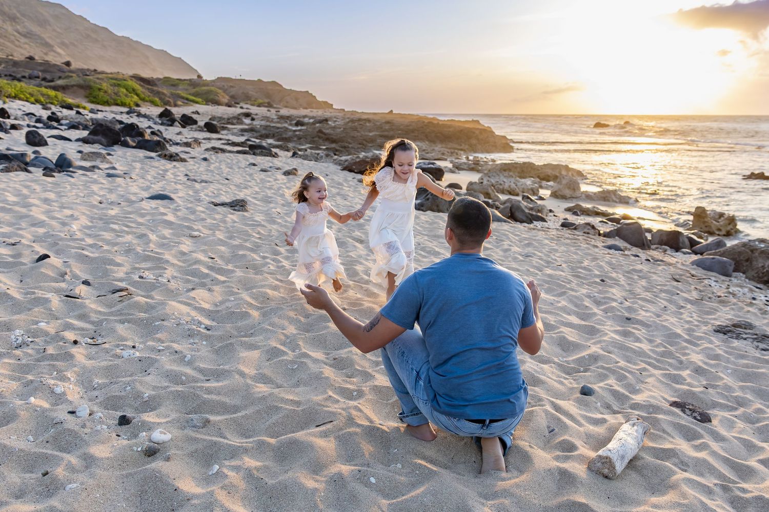 A family in white outfits enjoys a sunset photo session on a sandy beach with mountains and ocean in the background.