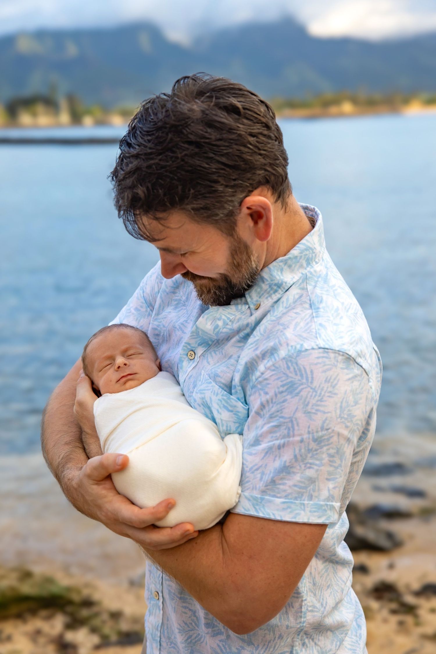 Tender moment between parent and sleeping newborn baby against ocean backdrop at sunset.