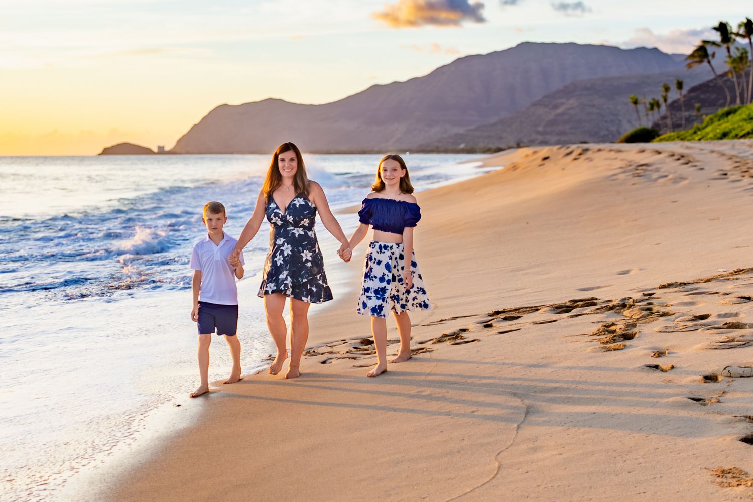 Three people walking together on a scenic beach at sunset with mountains and palm trees in view.