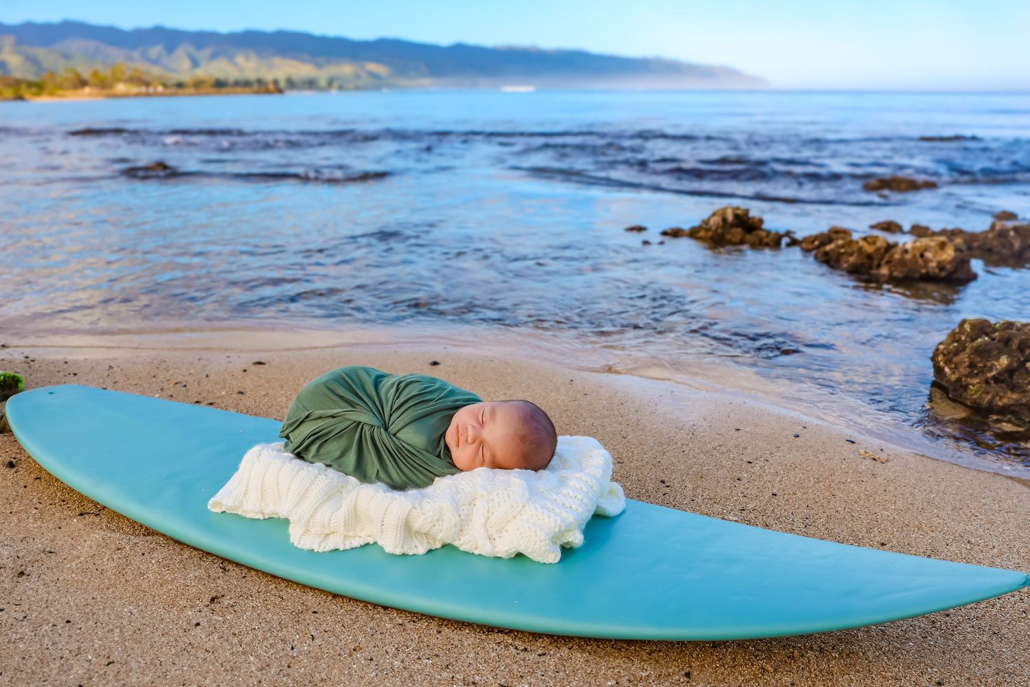 A sleeping baby lies on a blue surfboard at the beach with ocean and mountains in view.