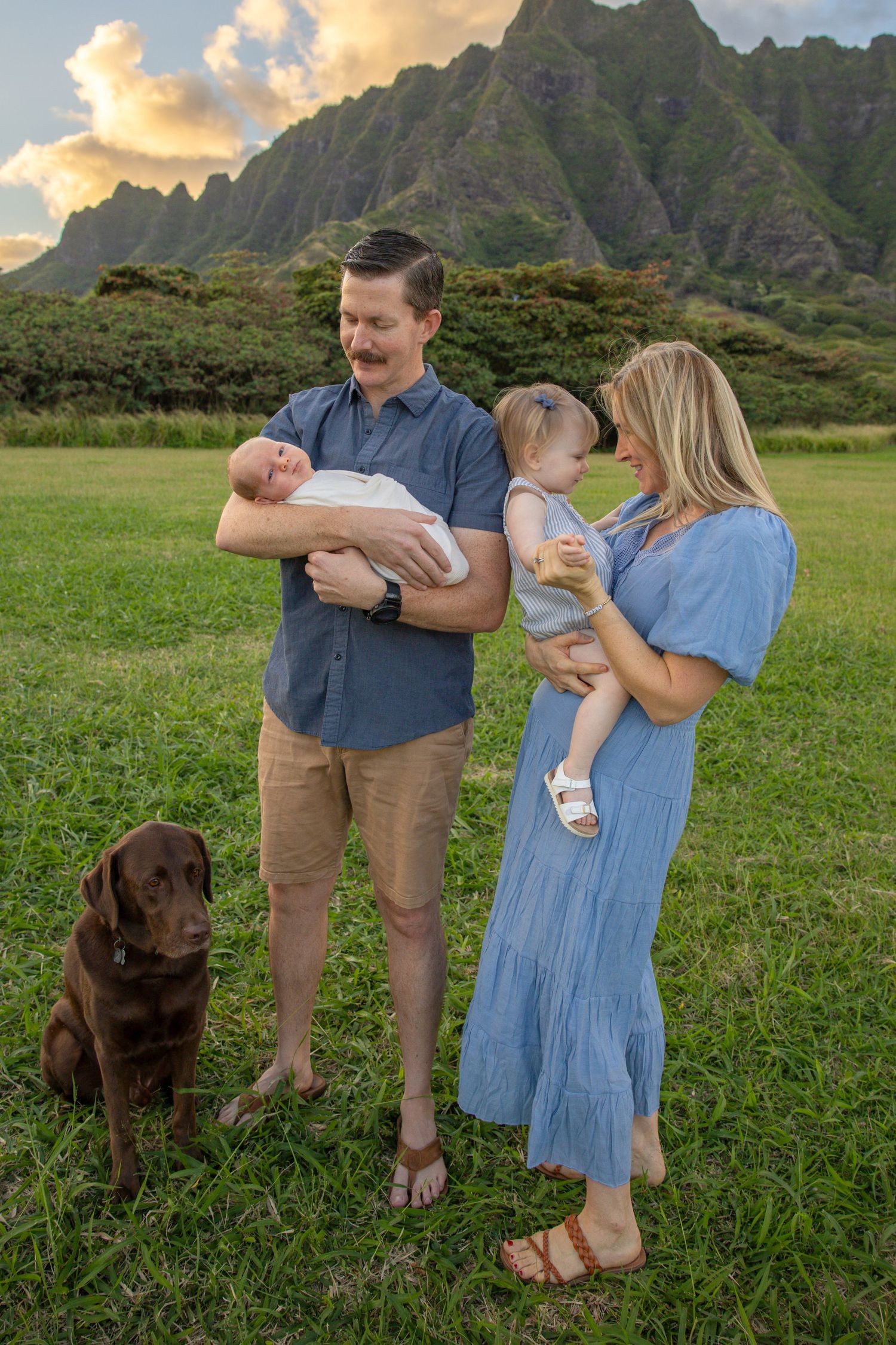 A family with a chocolate lab poses together on grass with dramatic mountains in the background.