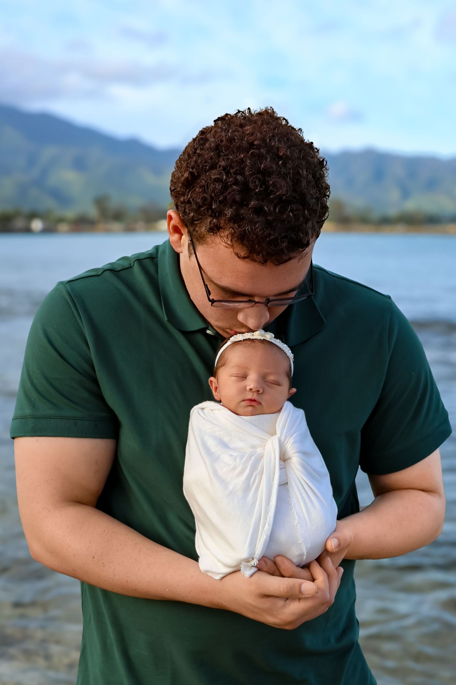 Parent in green polo shirt tenderly holding sleeping newborn wrapped in white against mountain backdrop.