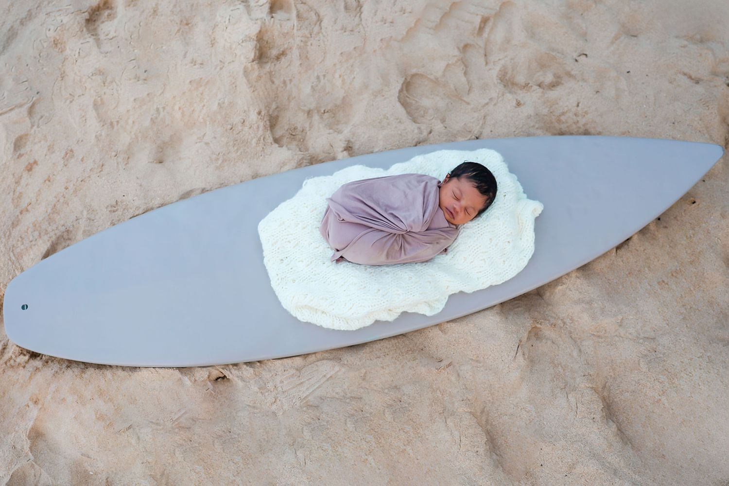 A baby wrapped in pink swaddle blanket sleeps peacefully on a surfboard in the sand.