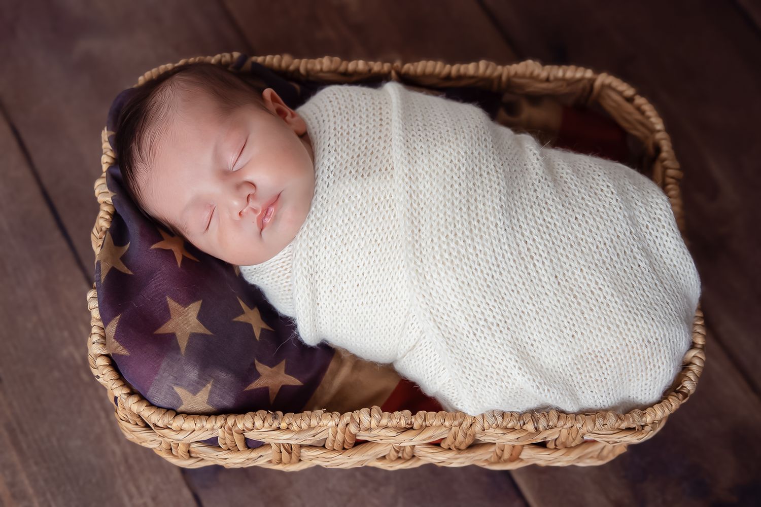 Sleeping newborn wrapped in white swaddle resting in woven basket with American flag.