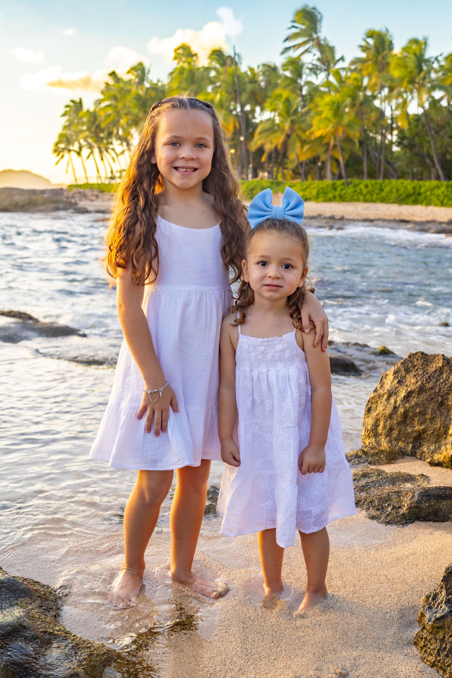 Two young girls in matching white dresses pose on a tropical beach at sunset.