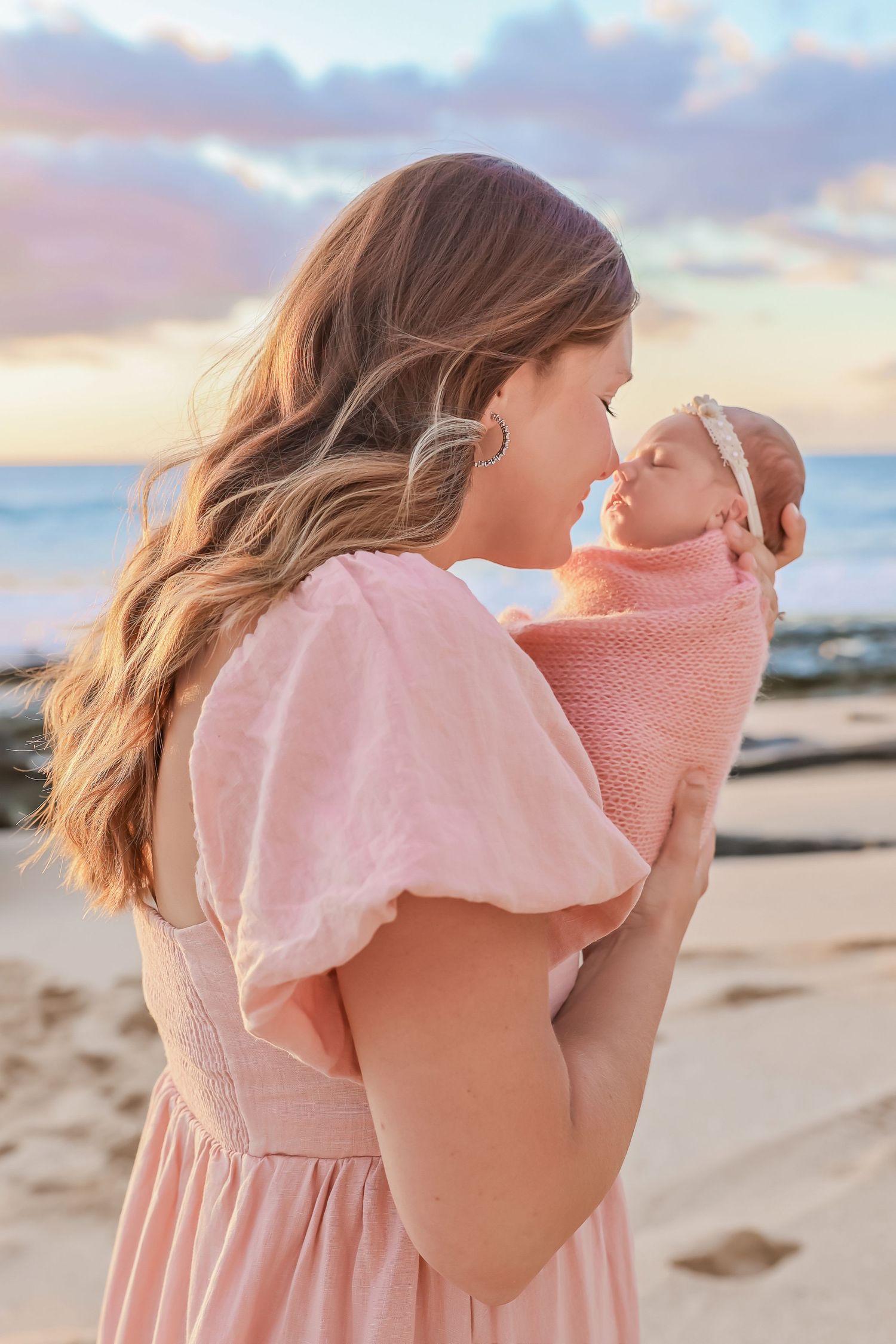 Gentle moment between parent and newborn baby wrapped in pink on beach at sunset.