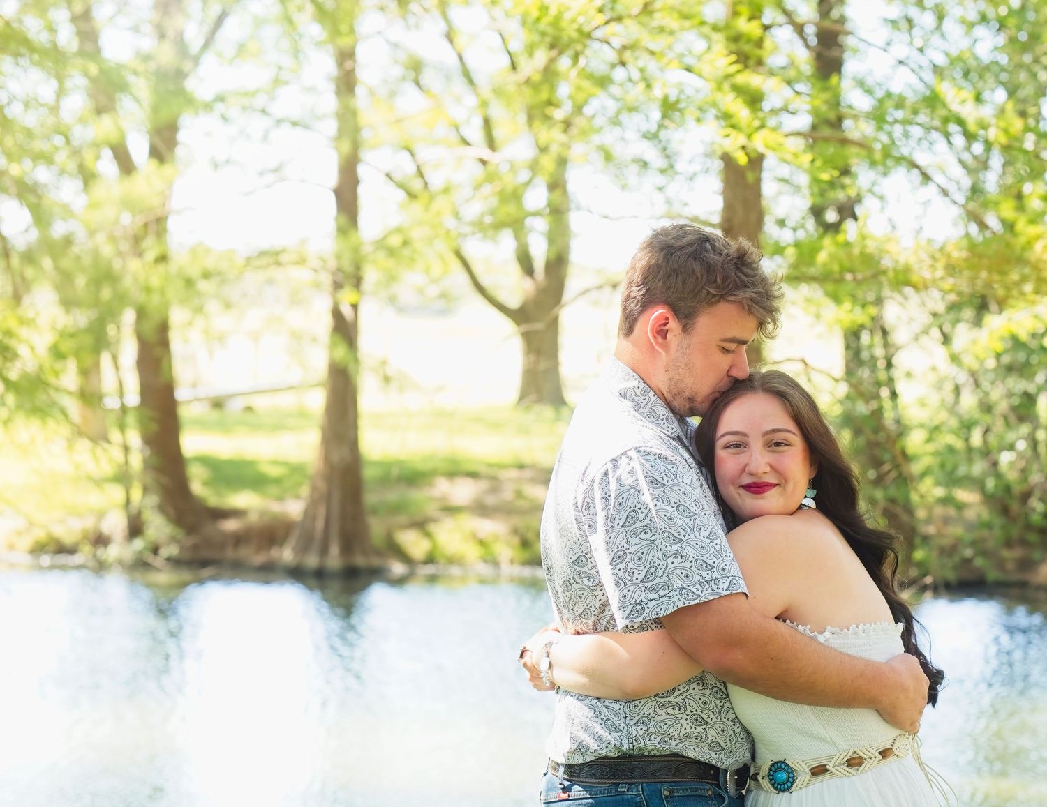 A couple shares a romantic embrace by a tranquil lake surrounded by sunlit trees in a natural setting.