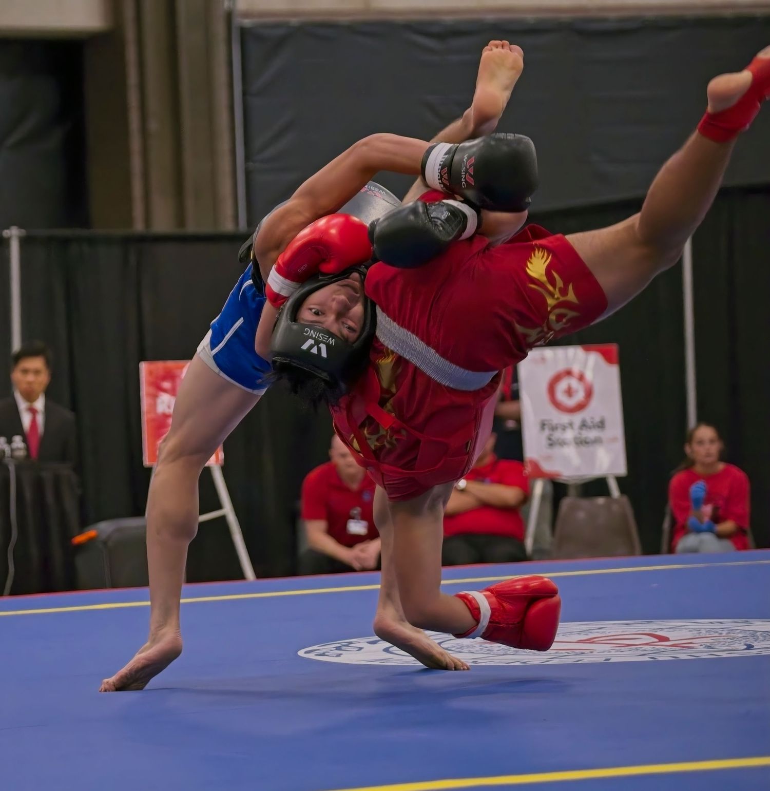 Two martial artists in red and blue uniforms perform dynamic kicks during a competitive match on a blue mat.
