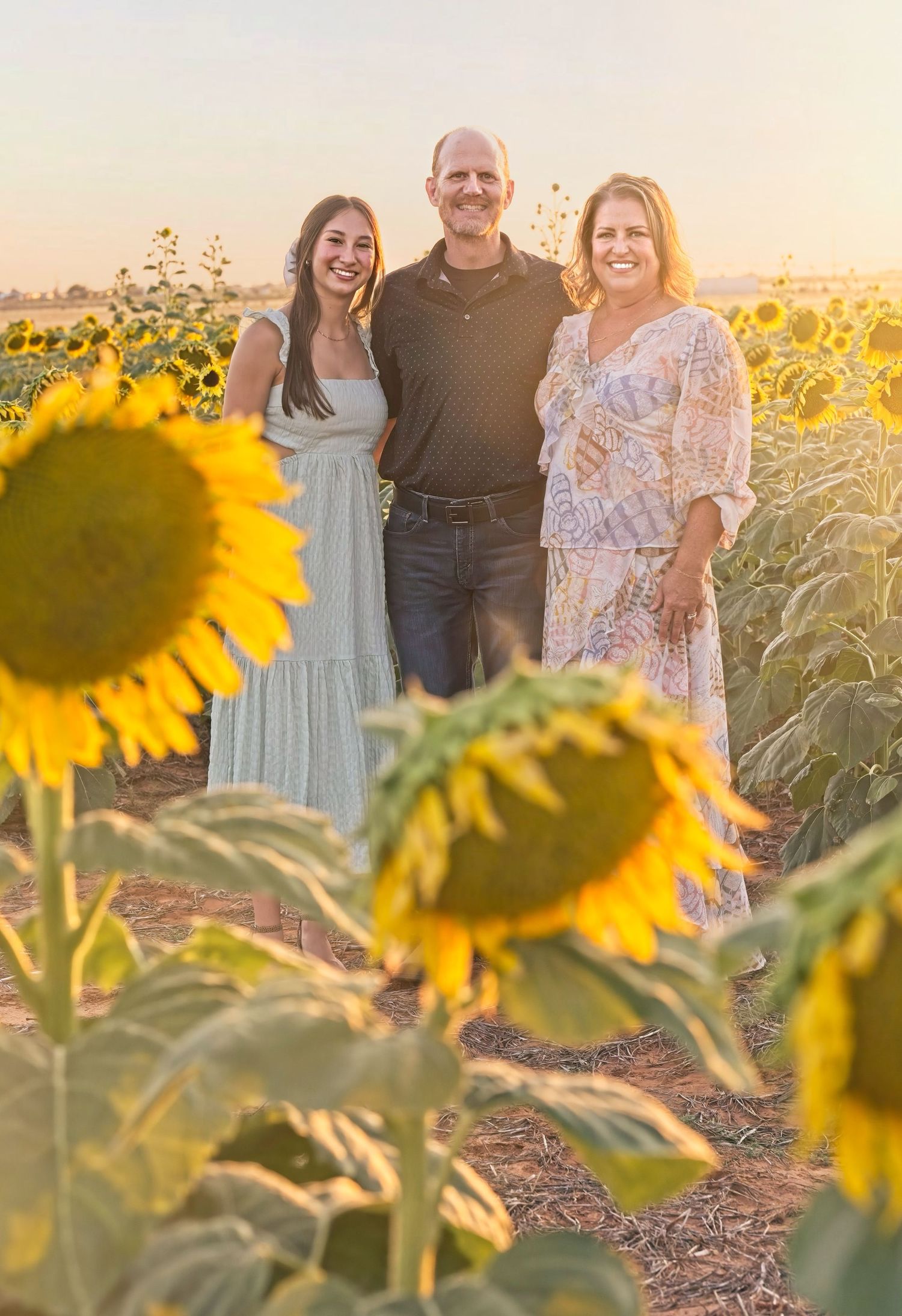 Three people stand smiling among tall sunflowers during a golden sunset.
