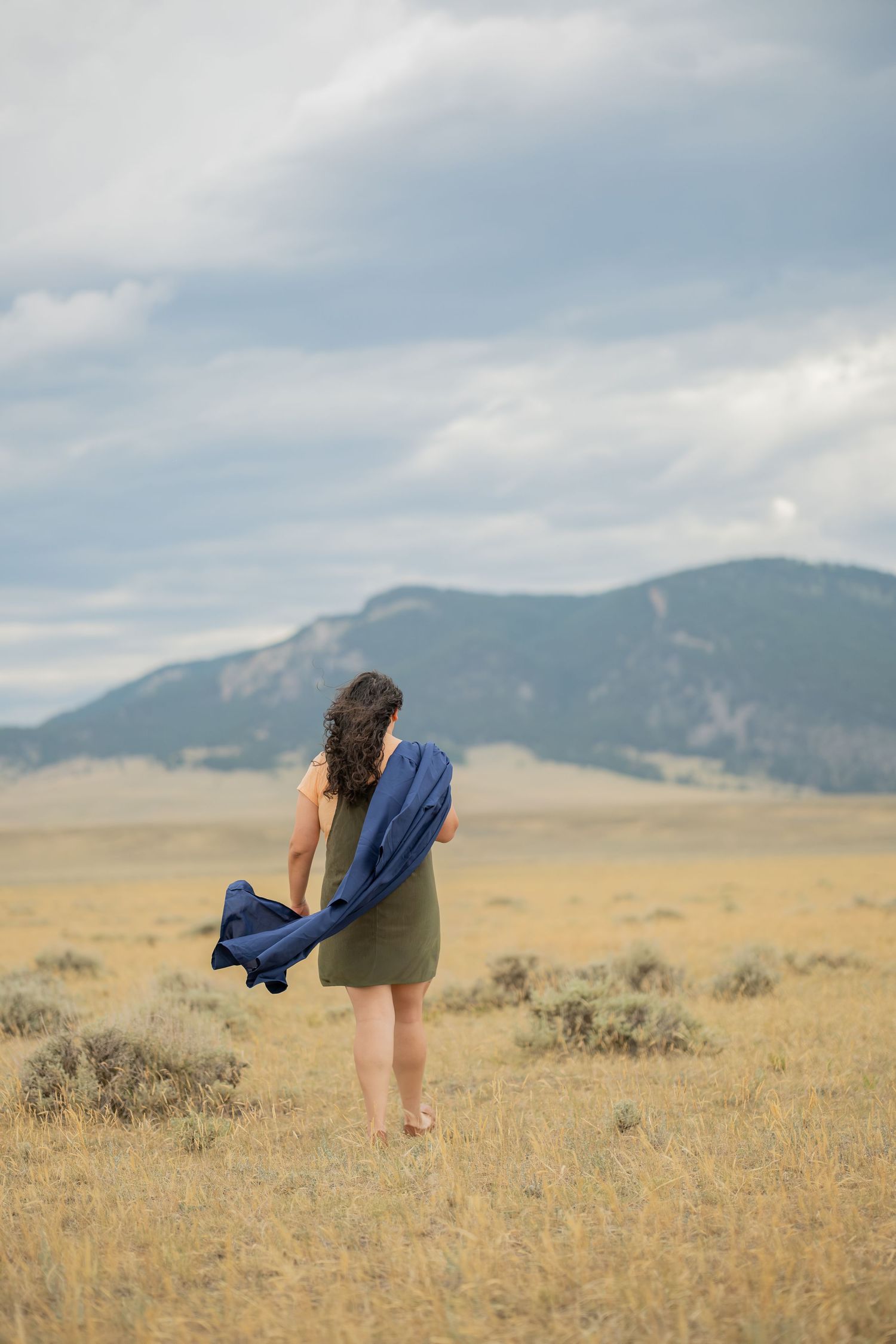 Woman in olive dress holding blue scarf walking through golden meadow with mountain backdrop under cloudy sky.