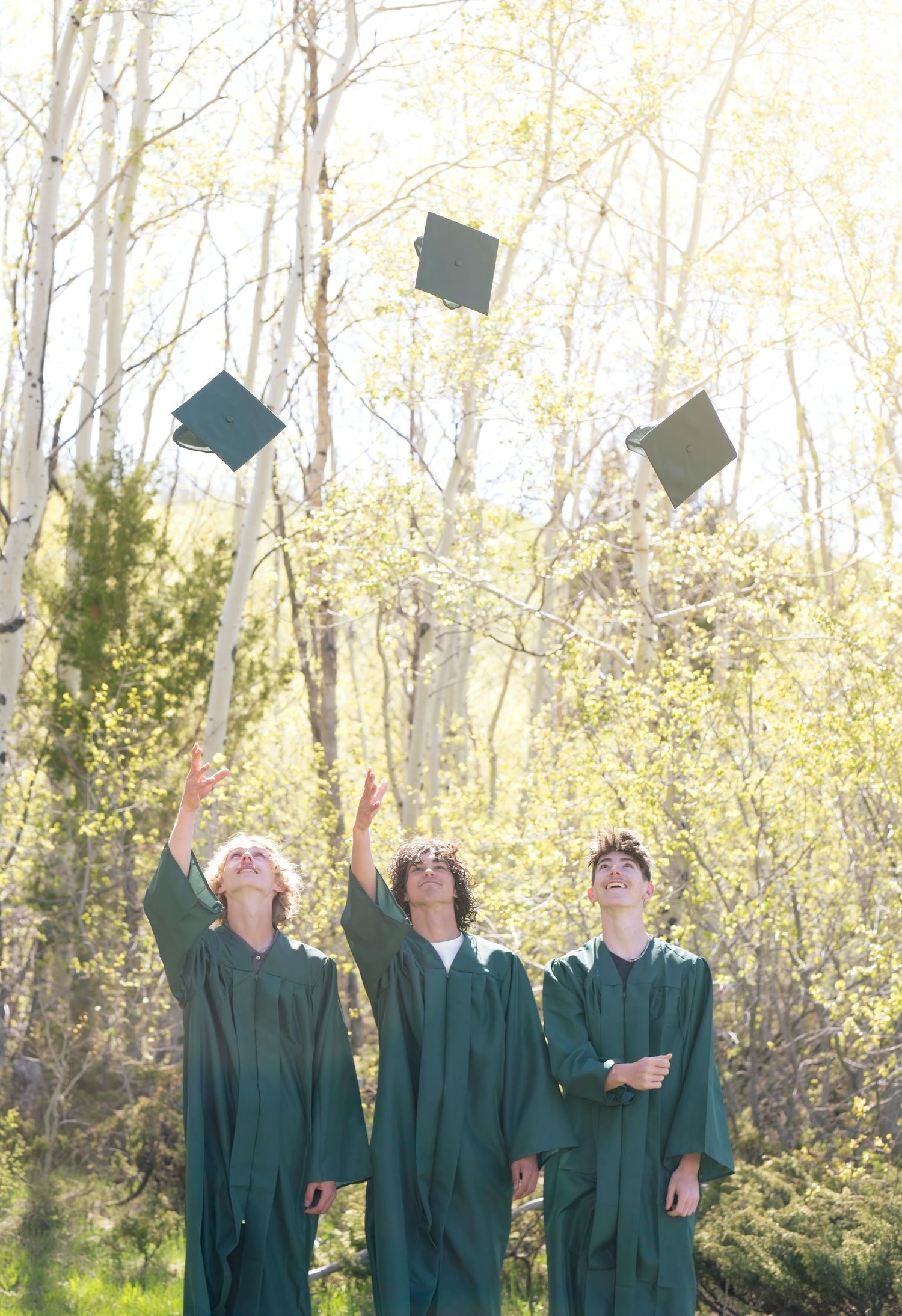 Group of graduates in green caps and gowns tossing their mortarboards into the air among spring trees.