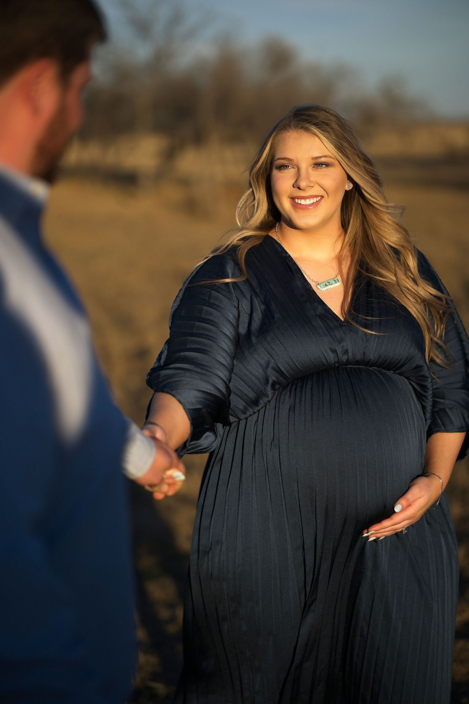 A couple holding hands during sunset for maternity photos, with a woman wearing a flowing black dress in warm light.