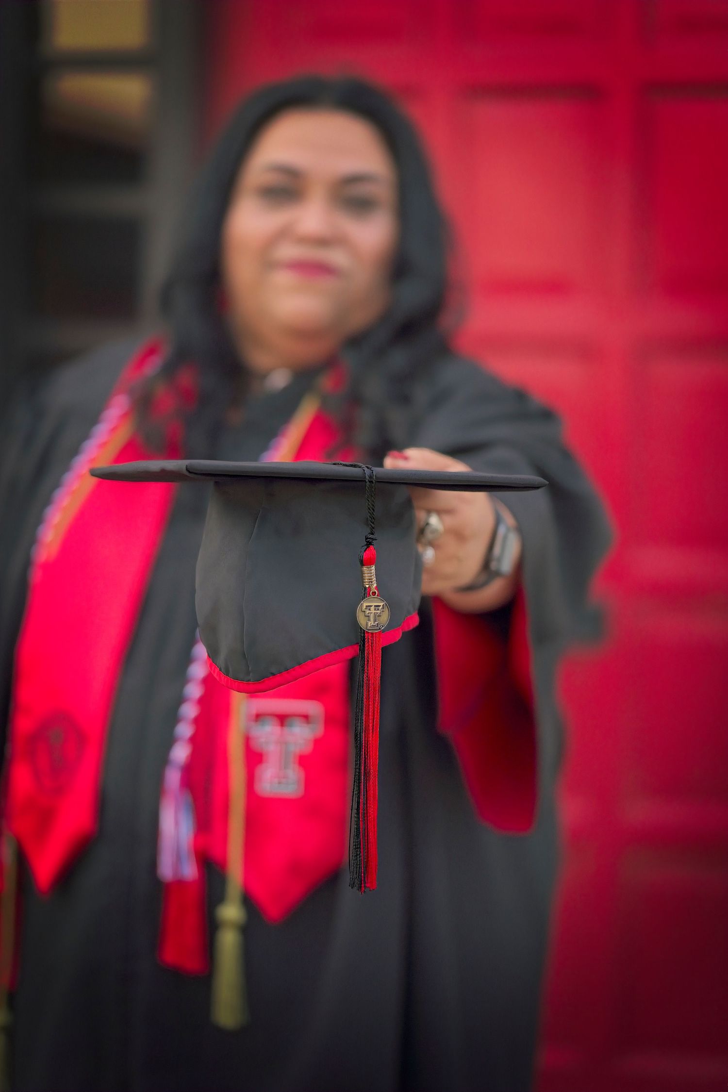 Graduate in cap and gown holding graduation cap with red stole against red door.
