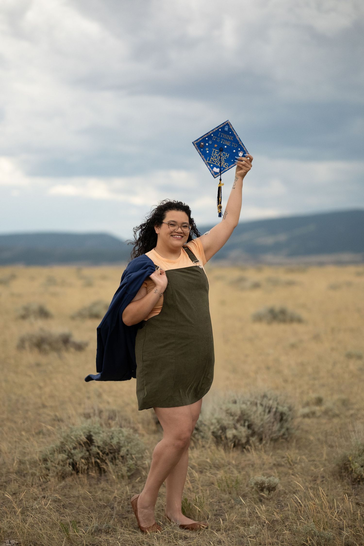 A smiling graduate holds up her decorated blue cap in a golden field, wearing a green dress and navy gown.