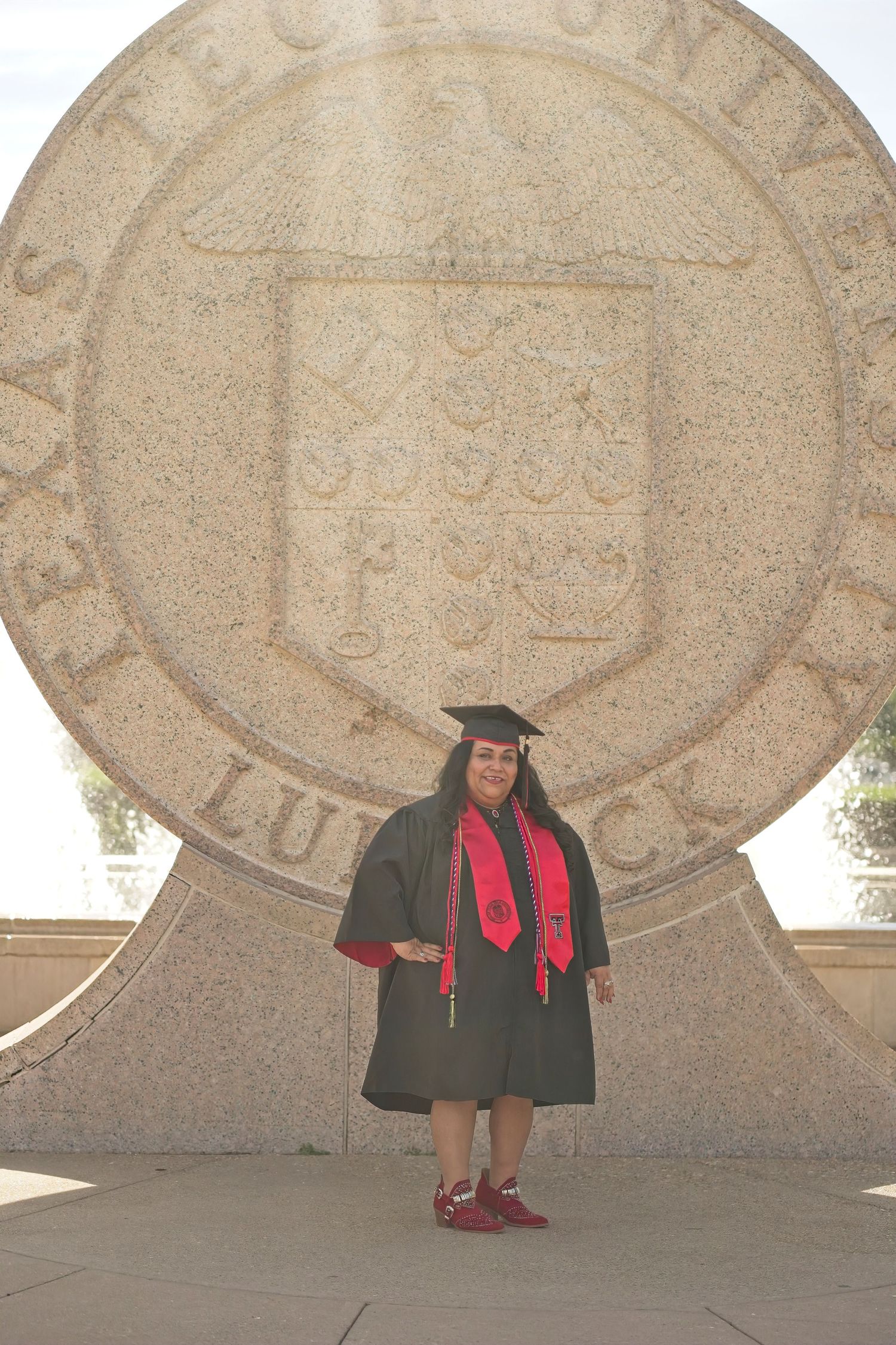 Graduate in academic regalia standing in front of large university seal monument.