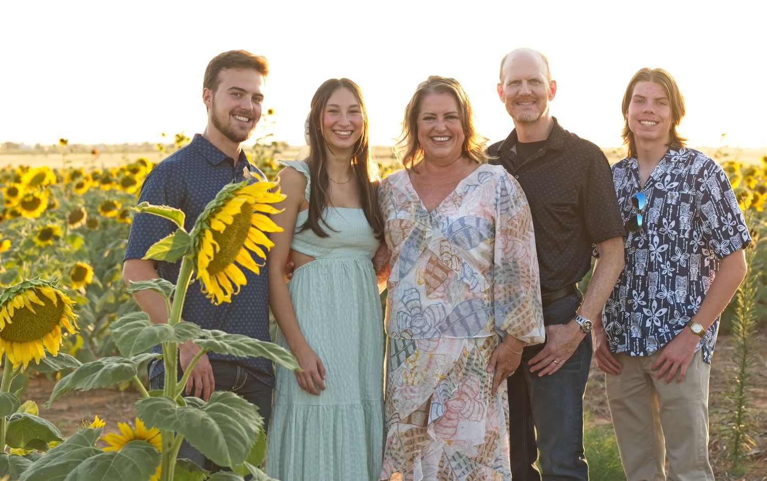 Five people pose casually together in a blooming sunflower field at sunset.