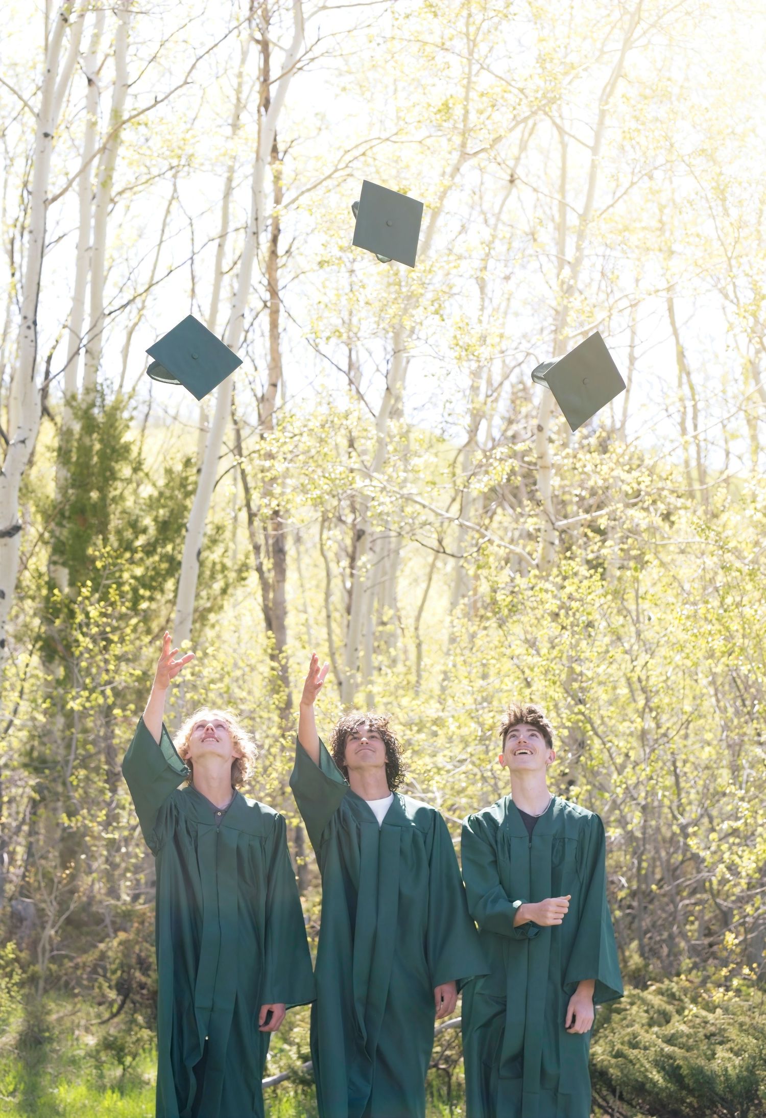 Graduates in dark green caps and gowns toss their mortarboards into the air during an outdoor graduation ceremony.