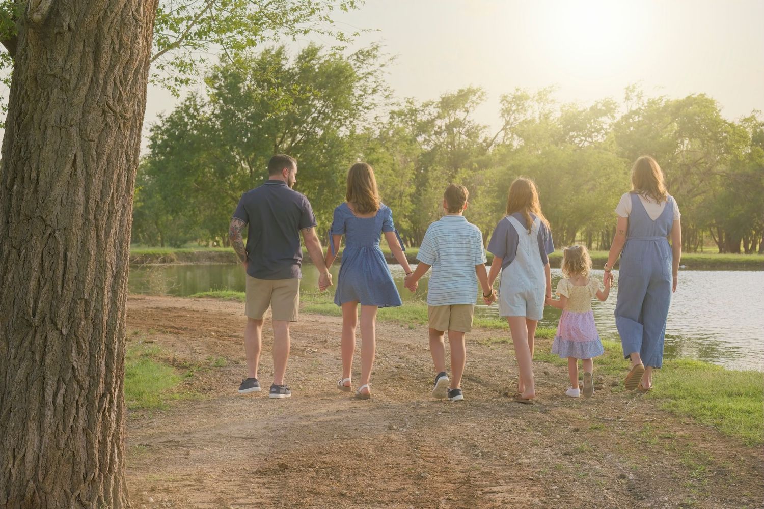 Group walking along lakeside path during golden hour beneath tall trees.
