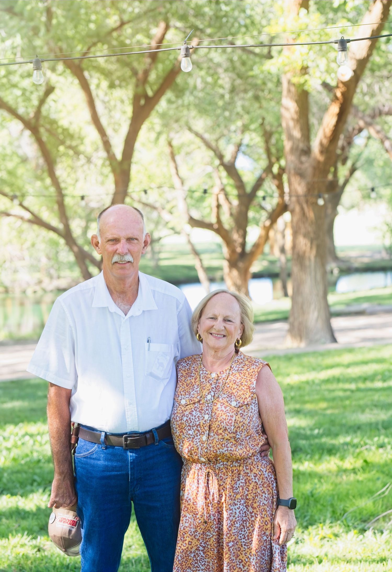 A couple in casual attire stands together outdoors in a sunlit park with lush green grass and trees in the background.