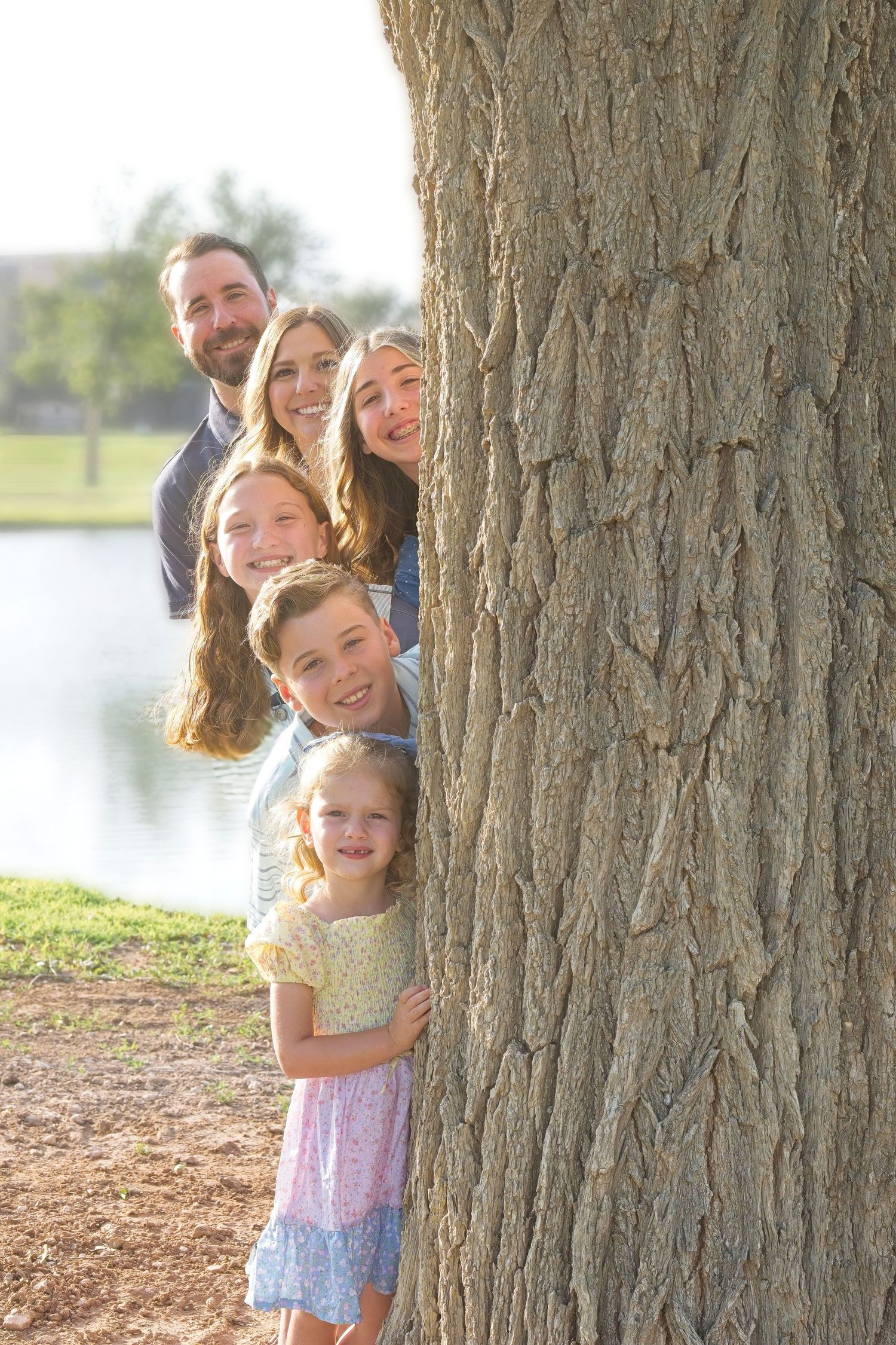 Playful scene with people peeking out from behind large tree trunks near a lake.
