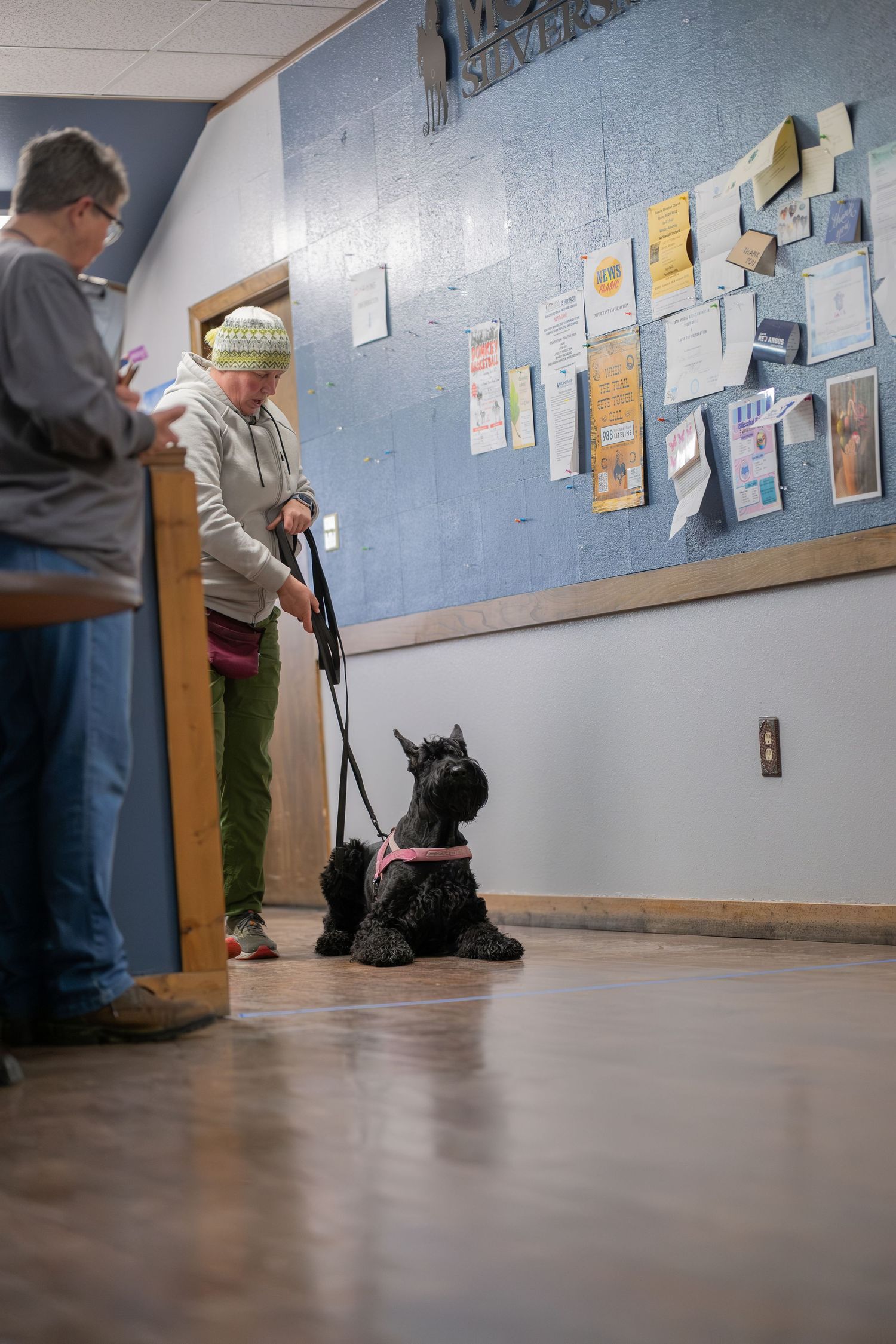 Woman in green hat walks black Scottish Terrier on pink leash inside a community center hallway.