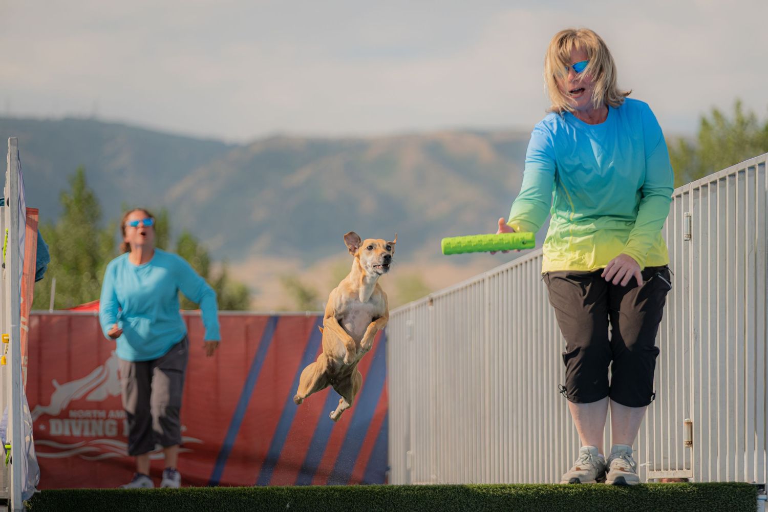 A woman in a colorful shirt catches a frisbee while a dog leaps mid-air at an outdoor disc dog competition.