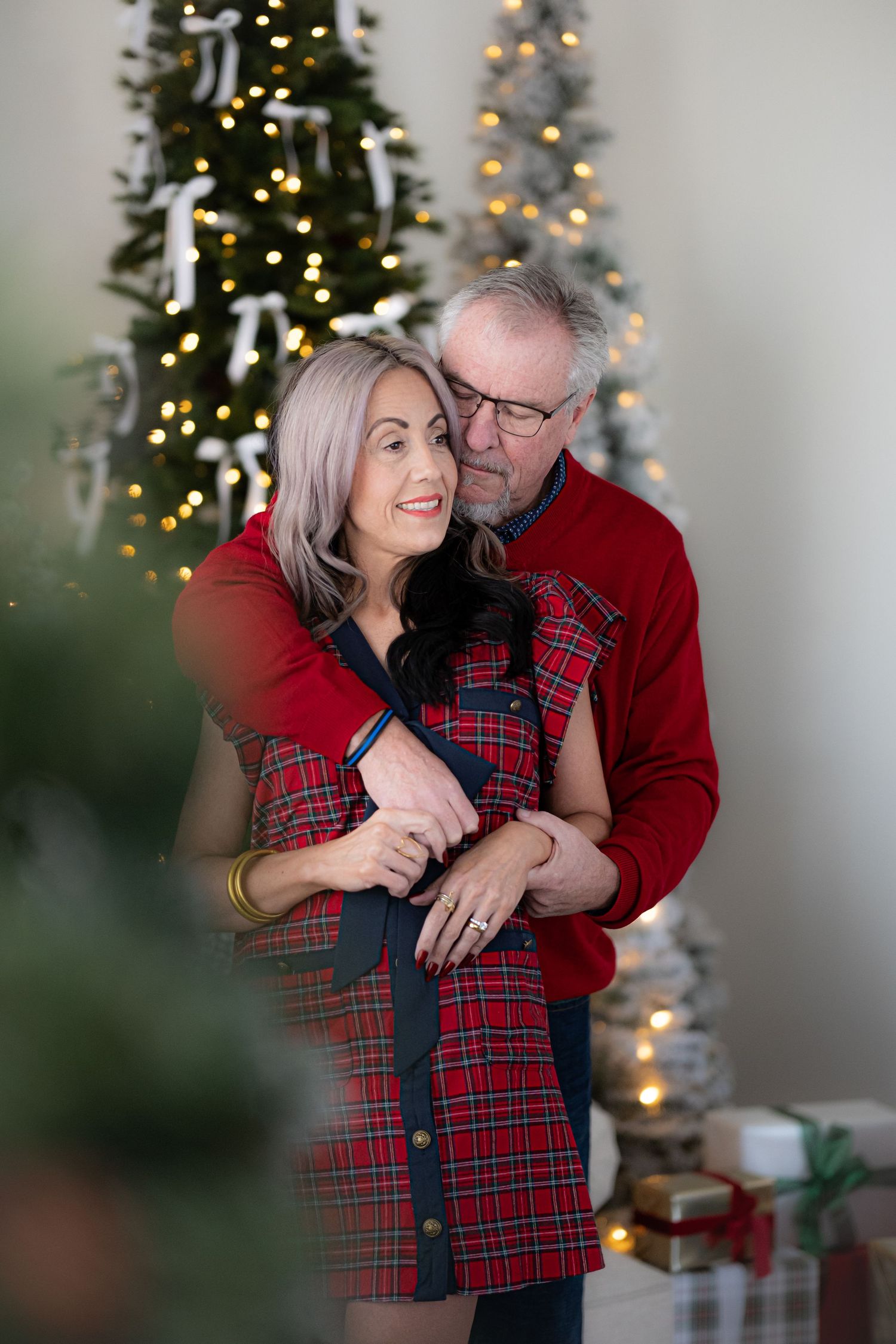 A couple in matching red plaid outfits share tender moments in front of a decorated Christmas tree.