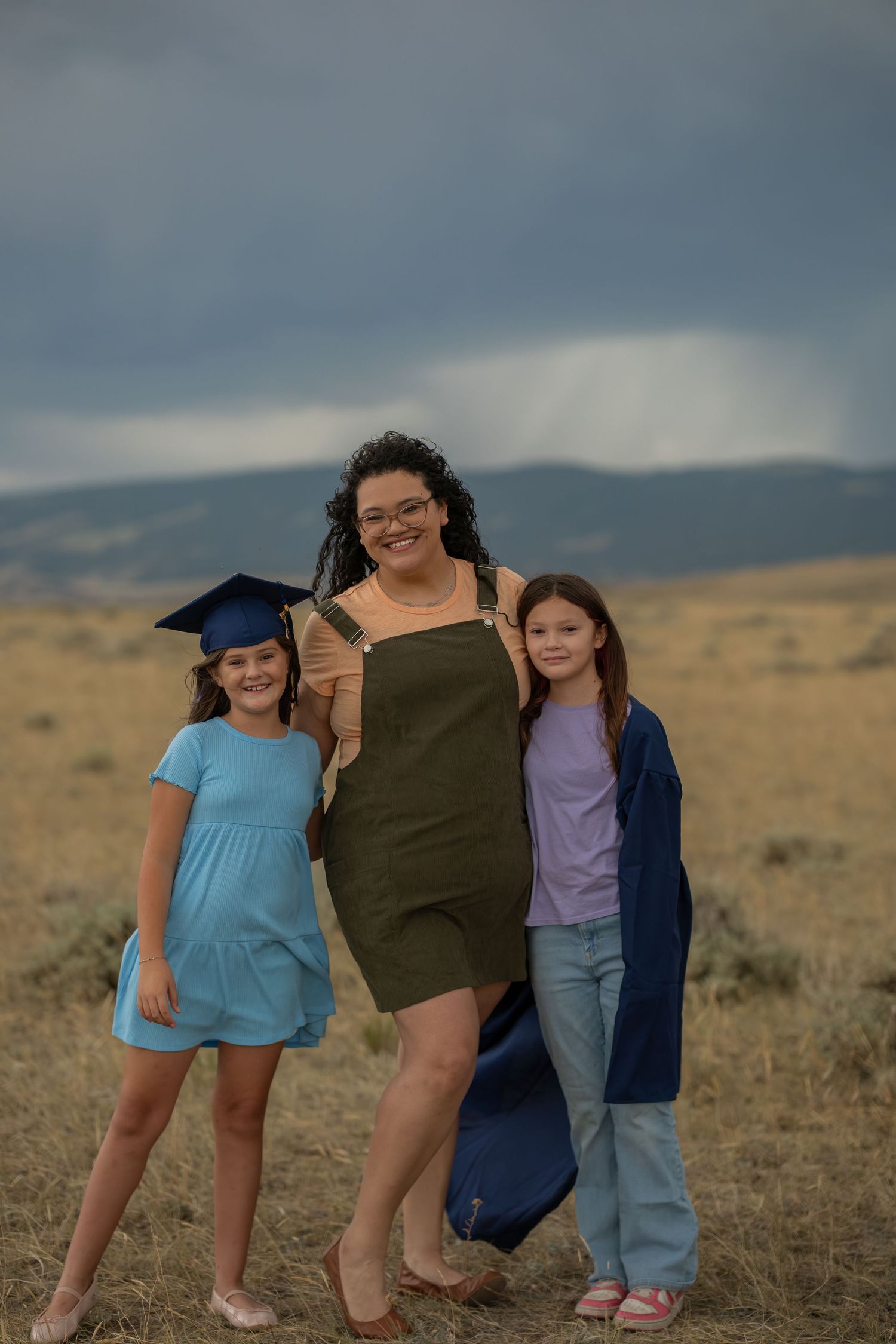 A woman and two girls pose outdoors, one girl wearing a graduation cap and gown, with stormy skies behind them.