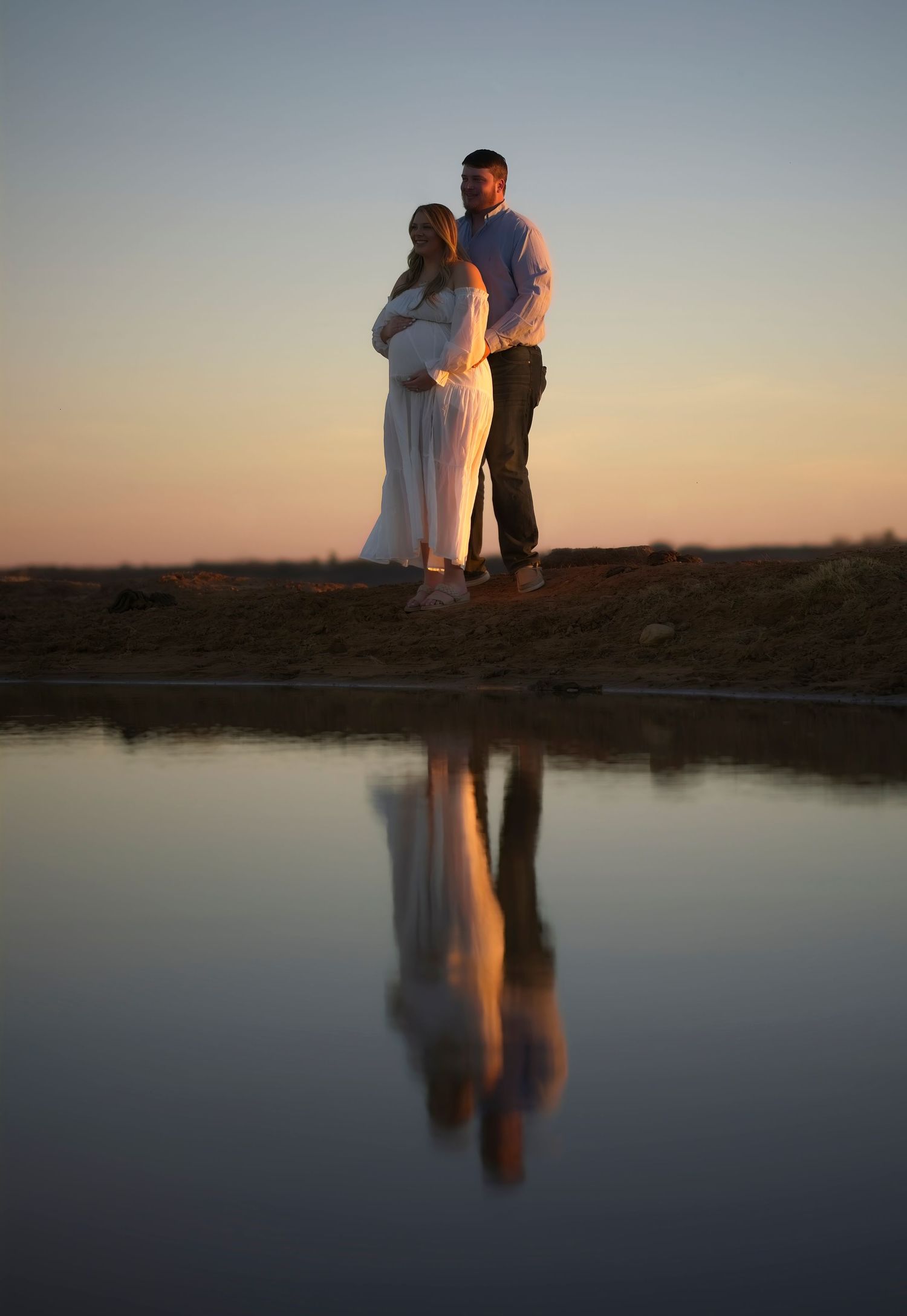 A couple stands silhouetted at sunset by a still pond creating a perfect mirror reflection in the calm water.