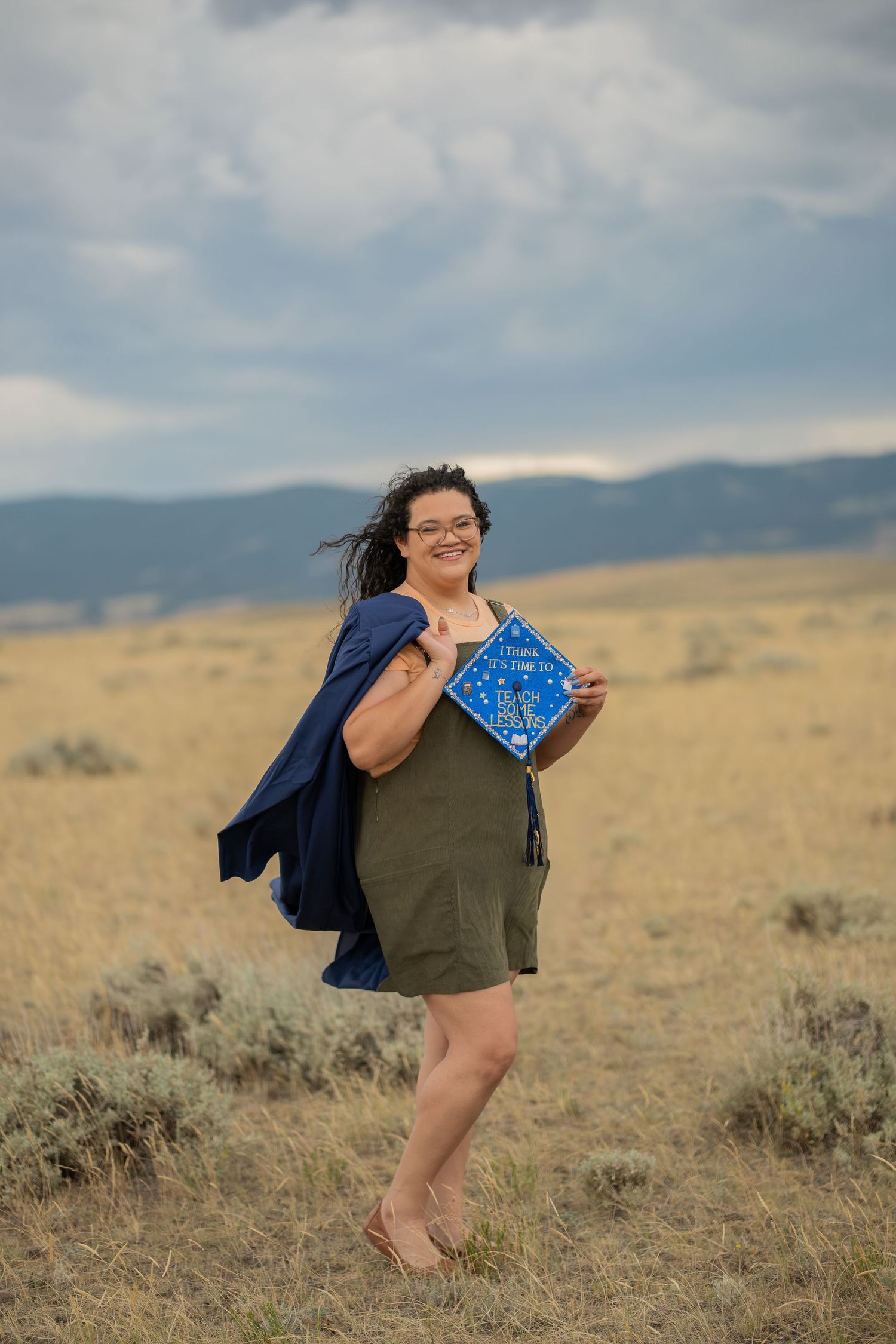 Woman in green romper holds blue graduation cap in golden field with mountain backdrop on cloudy day.