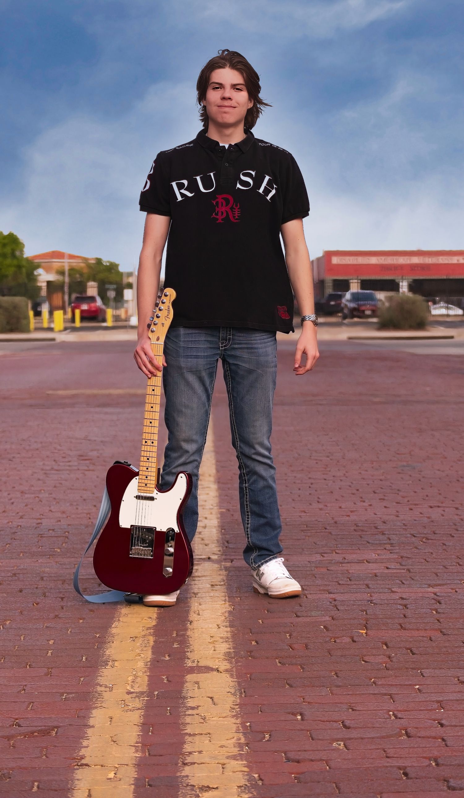 Musician poses with red Telecaster guitar on brick pavement.