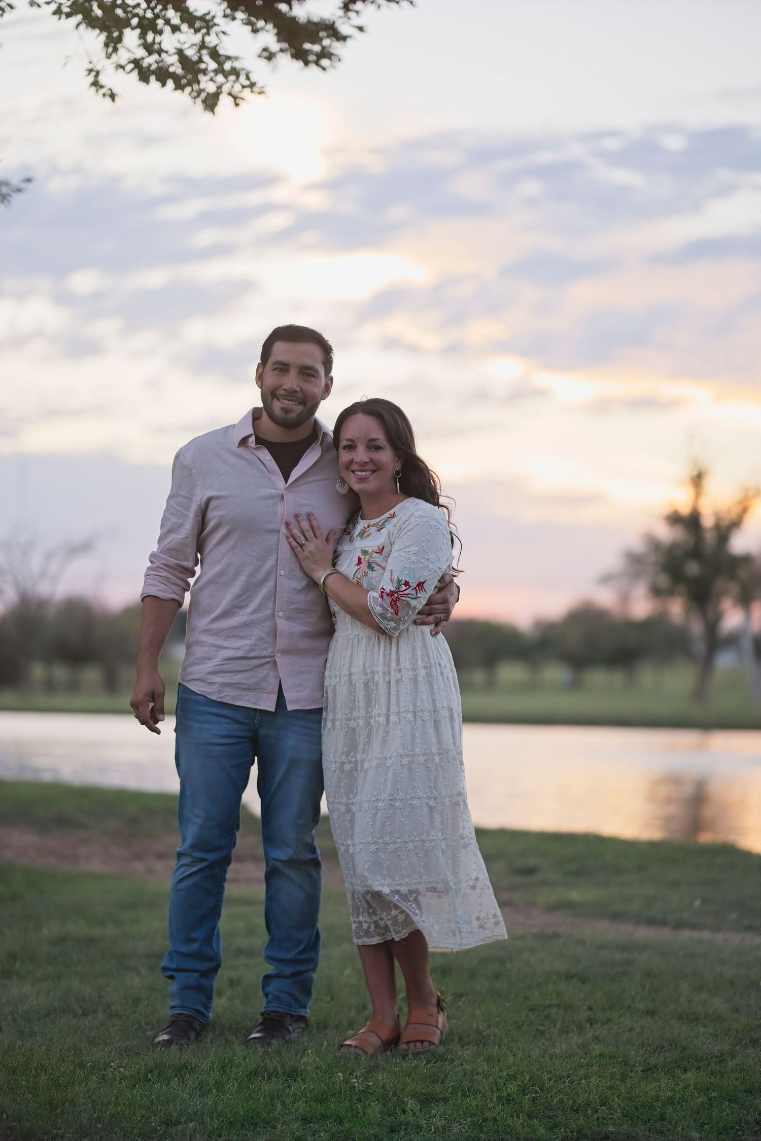A couple stands together on grassy field near water during sunset in romantic outdoor engagement photos.