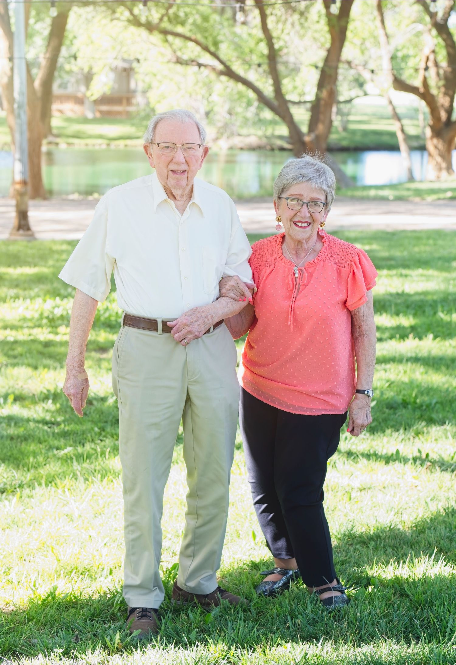 Senior couple walking and laughing together in a sunny park with trees and a pond in the background.