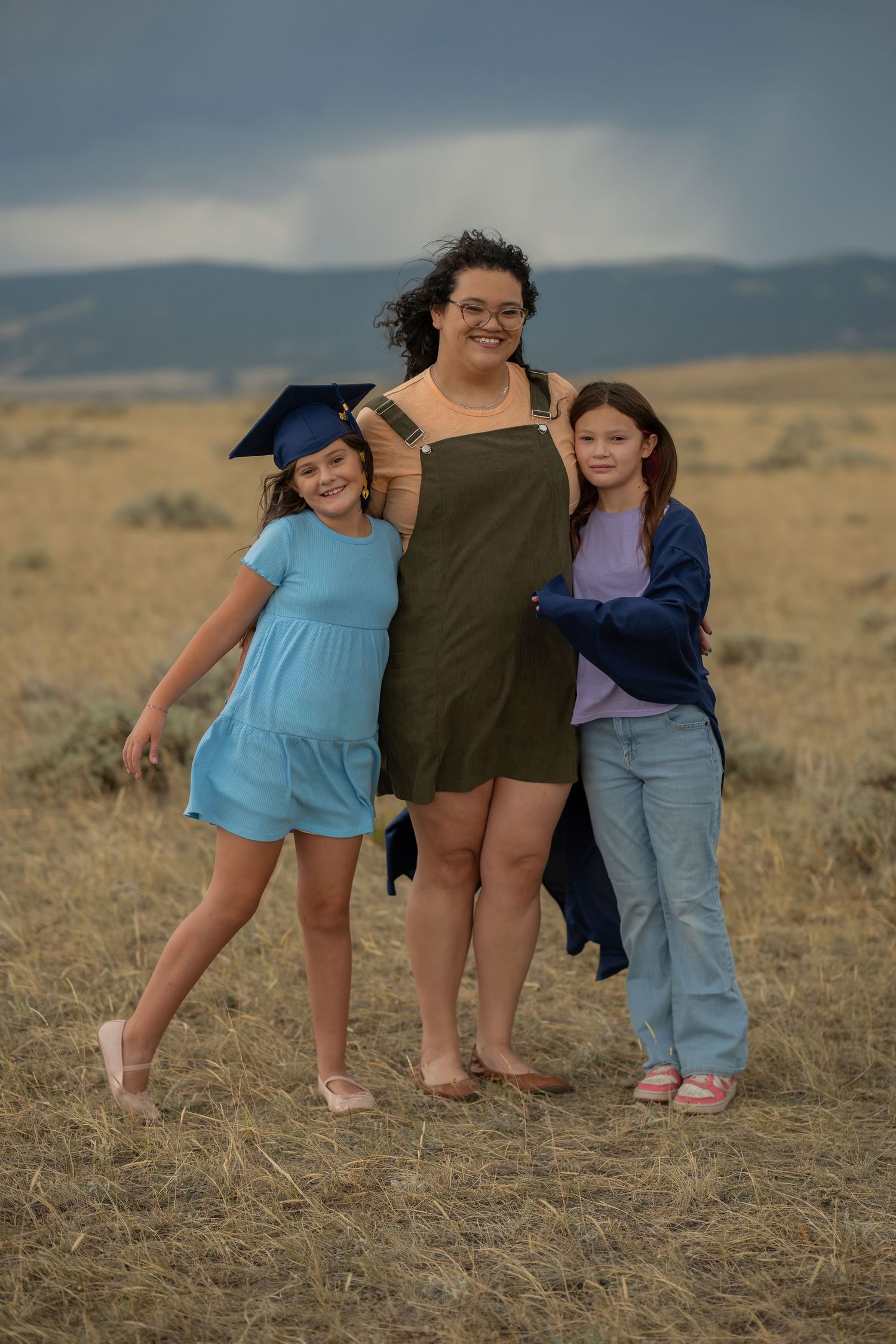 A woman in a green dress with two girls, one wearing a graduation cap, posing in a golden field under stormy skies.