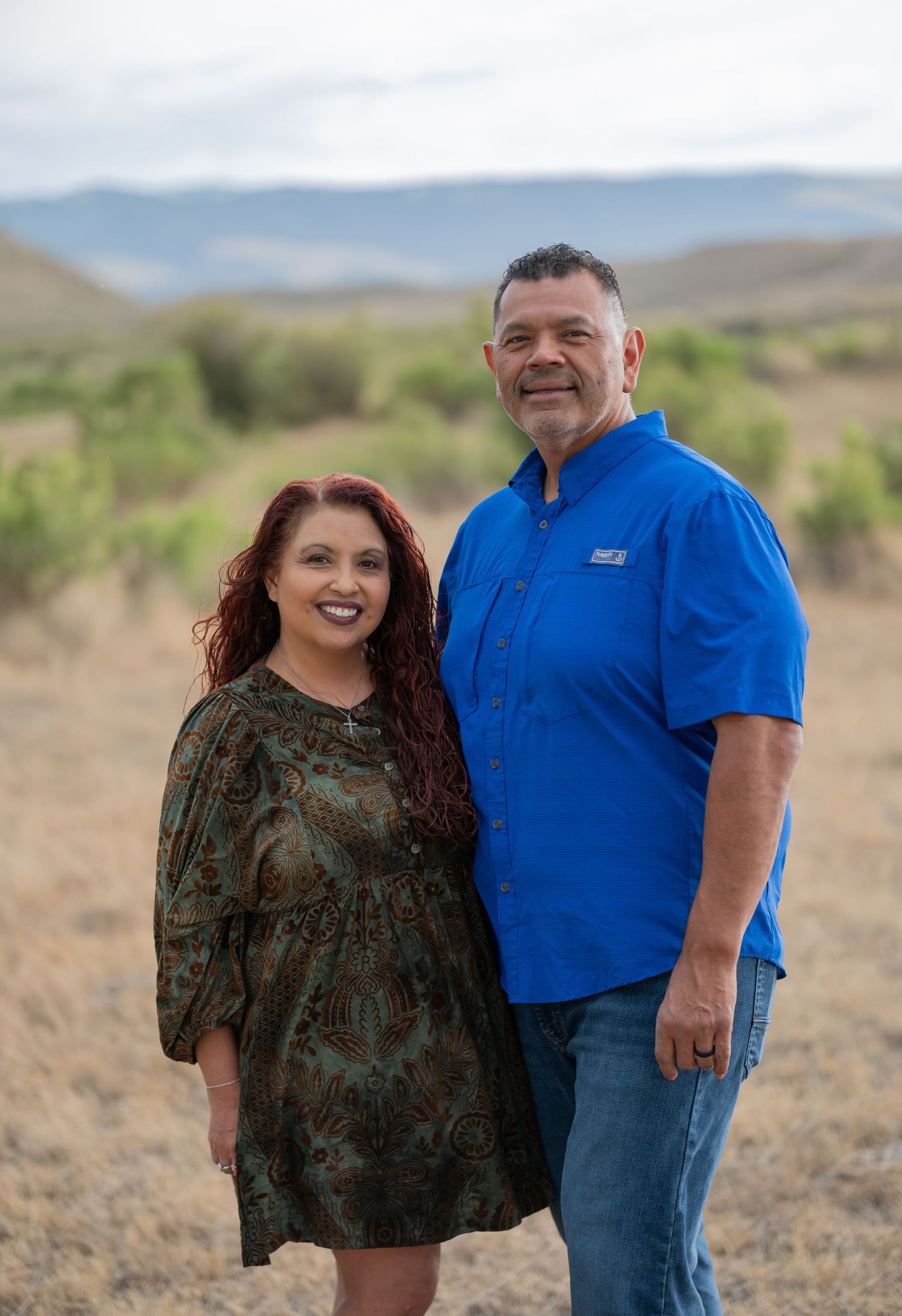 A smiling couple poses outdoors in a desert landscape, woman in floral dress and man in blue shirt.