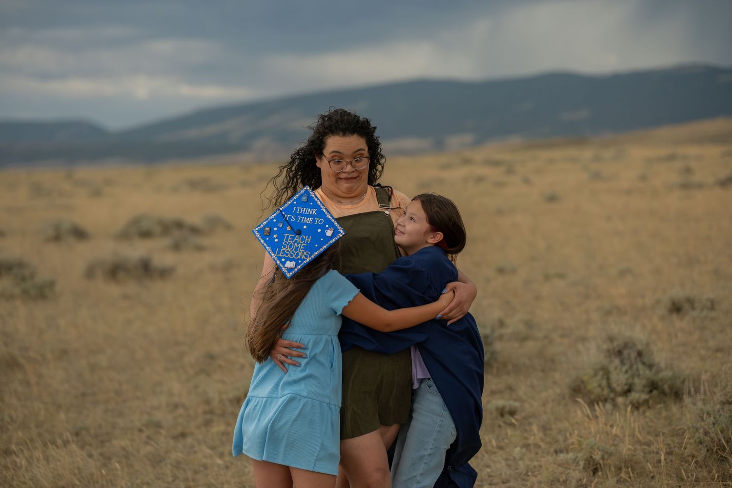 Graduate in cap hugs two people in golden field with mountain backdrop on cloudy day.